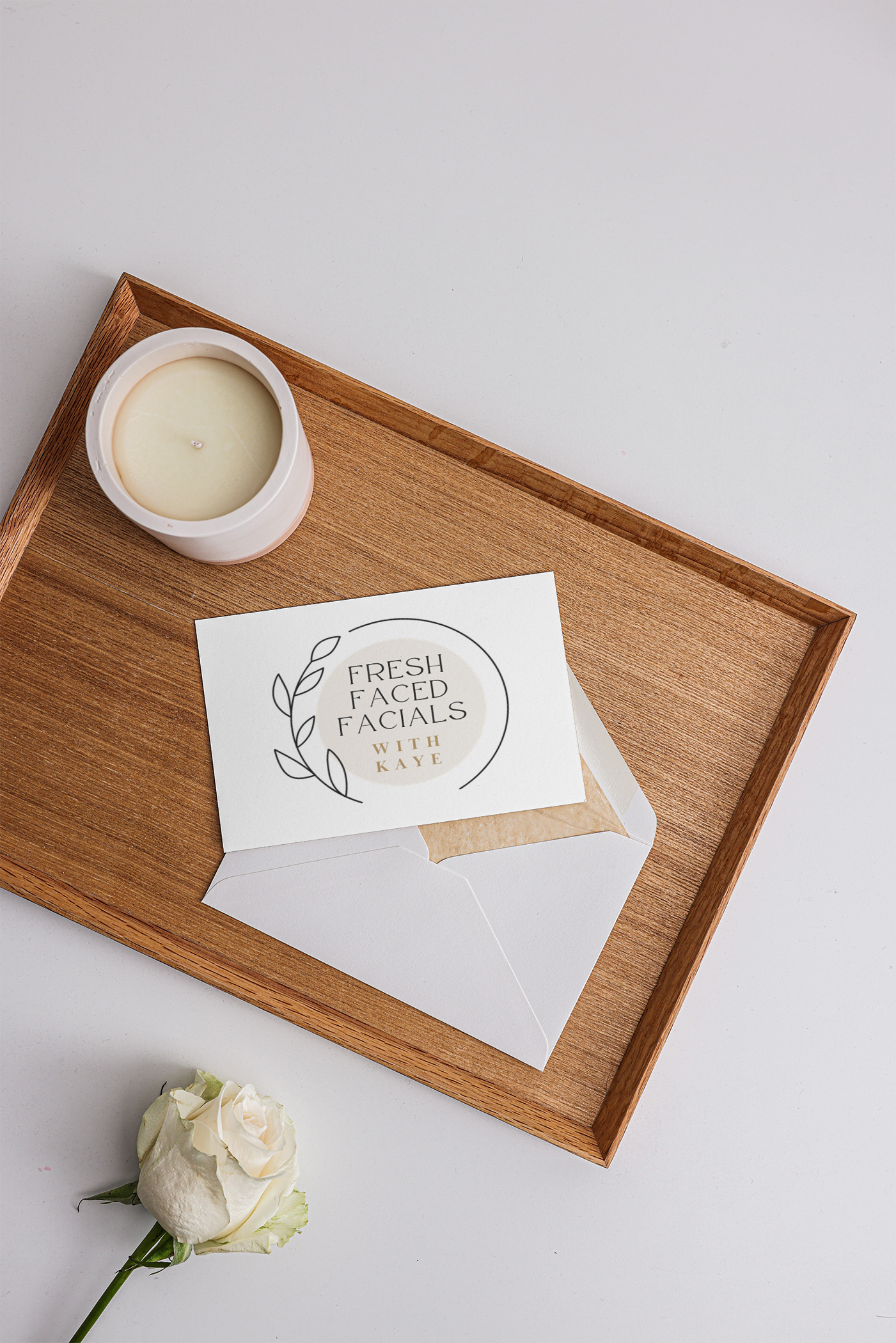 A white rose with a green stem lying on a white surface, with a wooden tray holding a white candle in a ceramic holder and a card labeled "Fresh Faced Facials with Kaye".