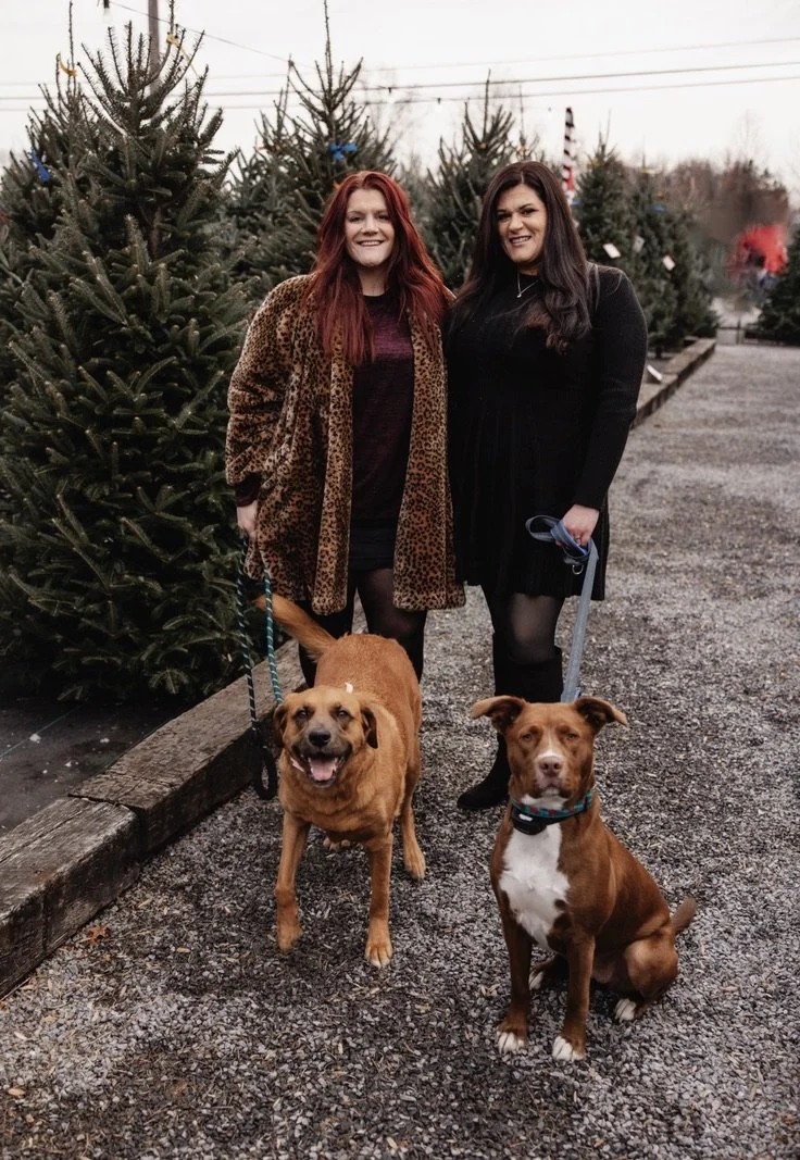 Two women standing outdoors on a gravel path with Christmas trees in the background. One woman has red hair and is wearing a leopard print coat, the other has dark hair and is dressed in black. Two dogs are sitting in front of them on leashes; one is smiling with a brown coat, the other is sitting calmly with a chestnut coat and white markings.