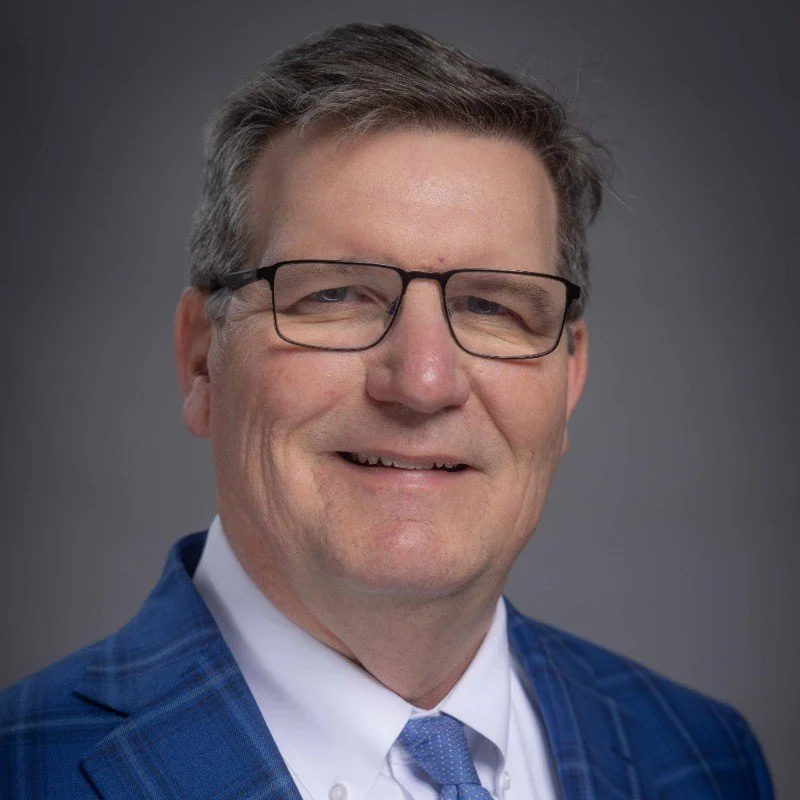 A middle-aged man wearing glasses, a white shirt, a light blue tie, and a blue blazer smiling against a gray background.