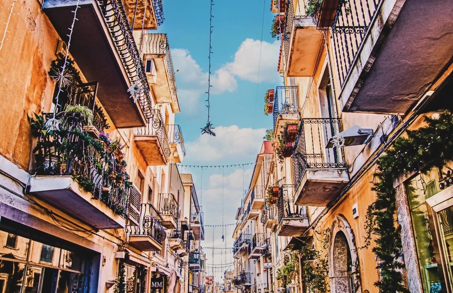 Narrow Mediterranean city street lined with balconies, potted plants, wrought-iron railings, and festive string lights under a blue sky