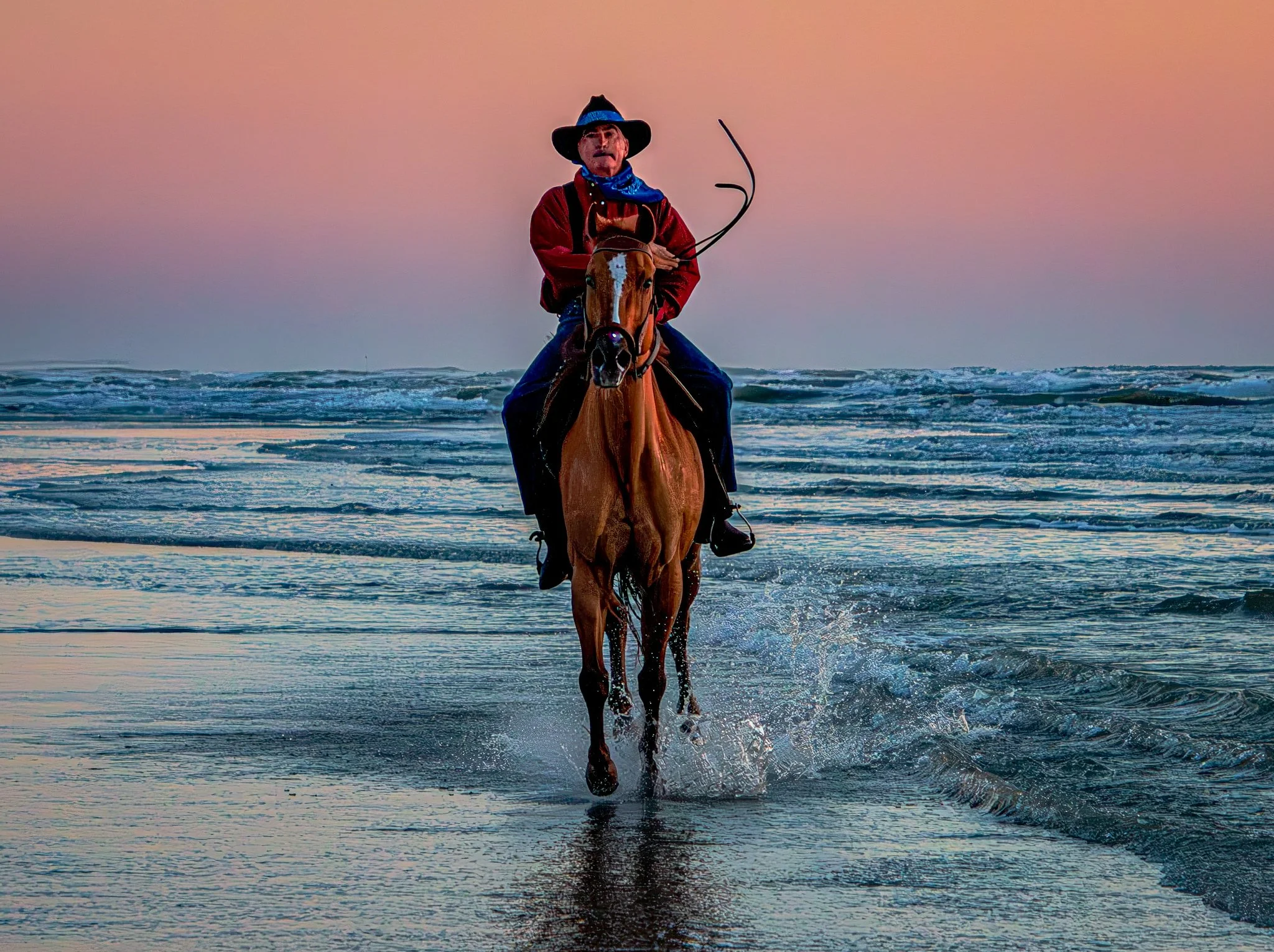 Person riding a horse along the shoreline at sunset, with waves splashing around the horse's legs and a pastel-colored sky in the background.