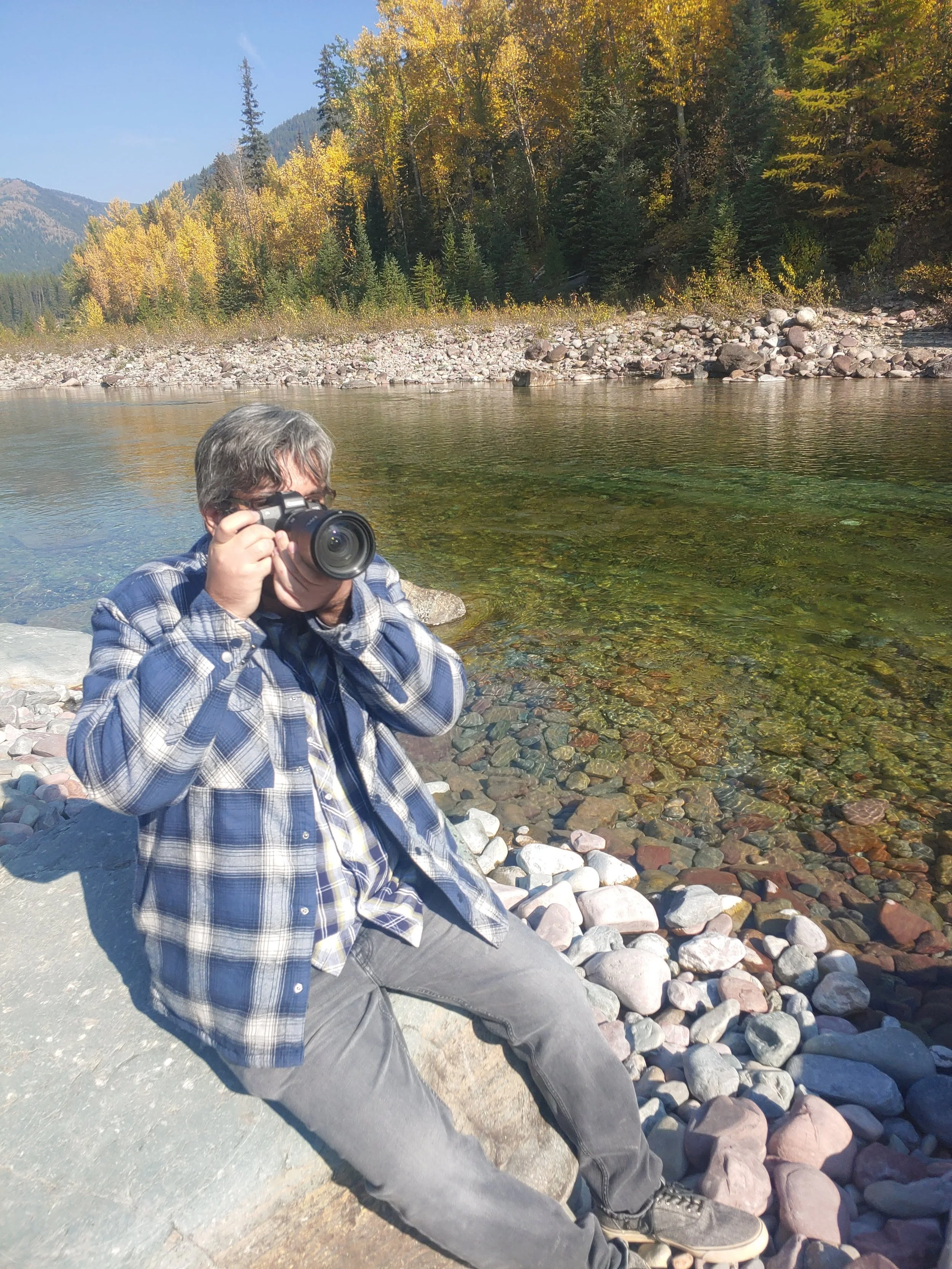 Photo of photographer Garrett Chinn holding a camera while sitting on Rocks by a River.