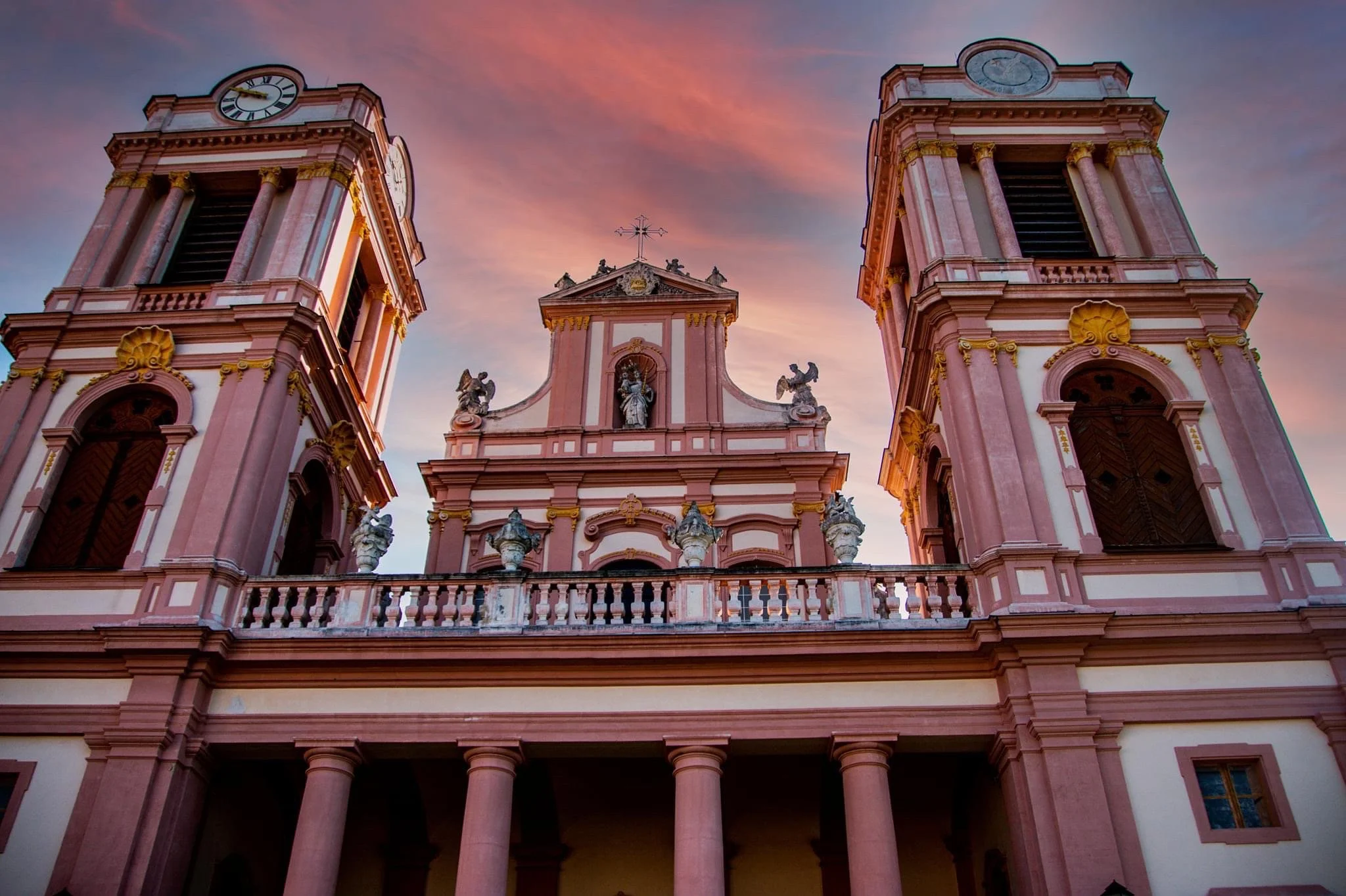 Ornate pink Baroque church facade with twin clock towers, statues, columns, and gold detailing at sunset