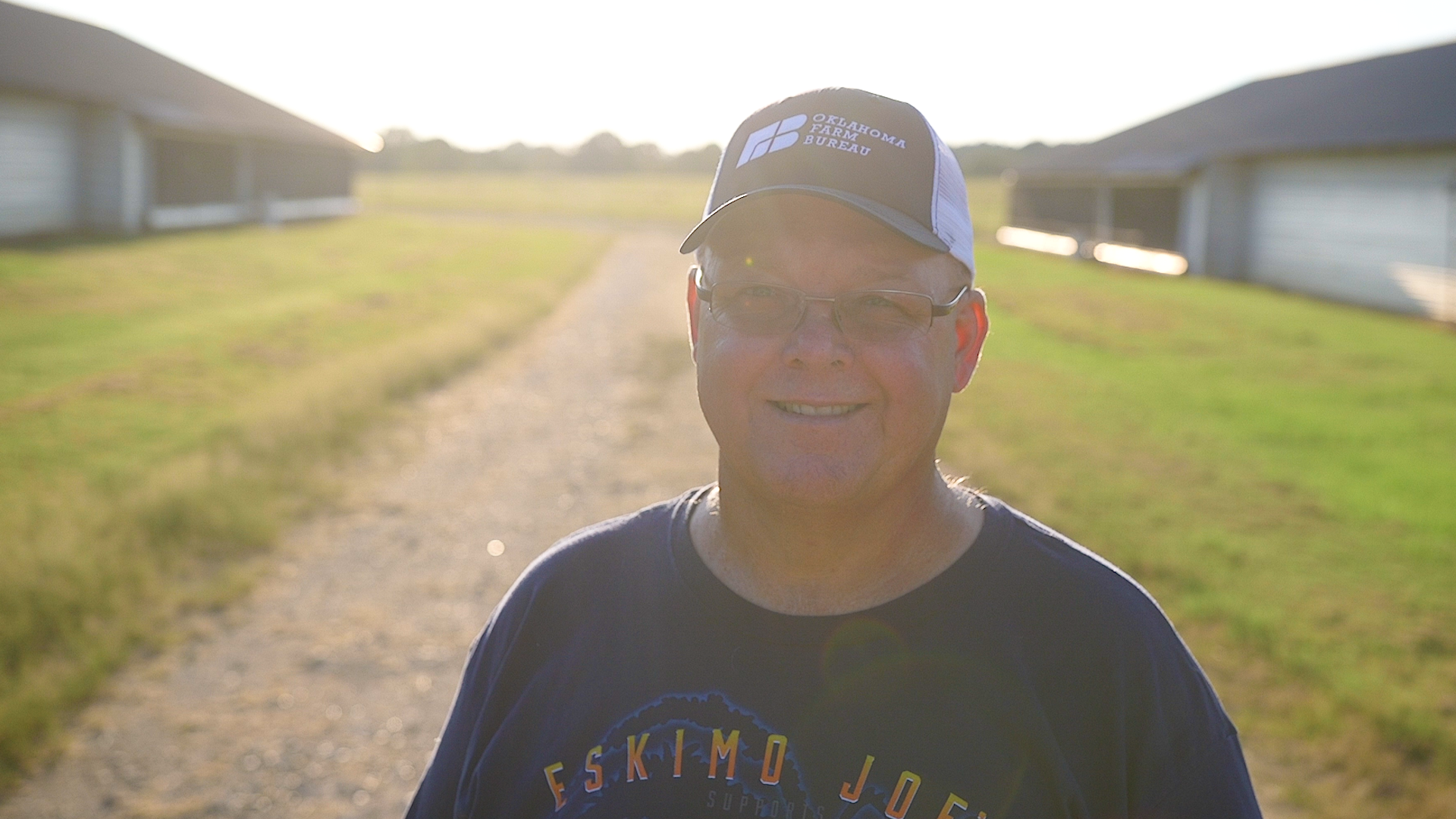 Smiling man wearing glasses, a baseball cap, and a T-shirt, standing outdoors on a dirt path with houses and green grass in the background, backlit by the setting sun.