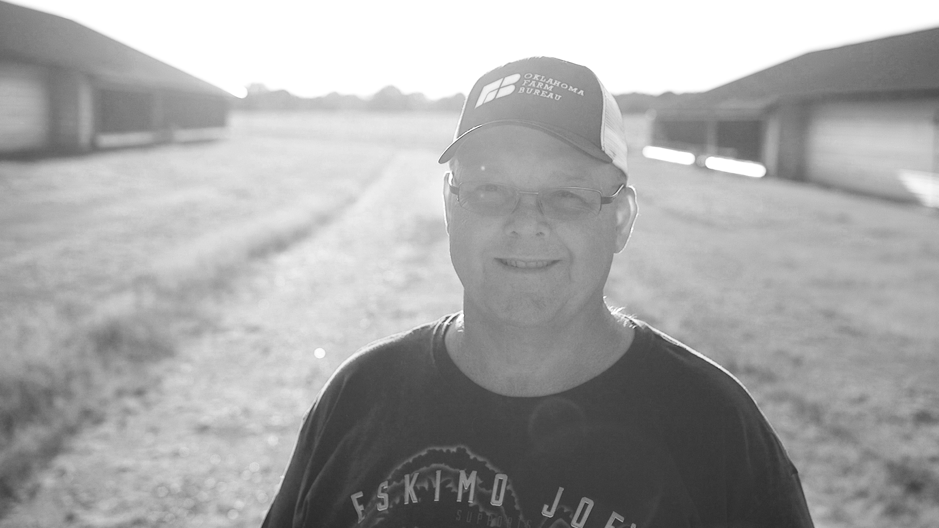 A smiling man wearing glasses and a baseball cap with Oklahoma Farm Bureau logo, outdoors on a farm, with barns and a gravel path in the background, in black and white.