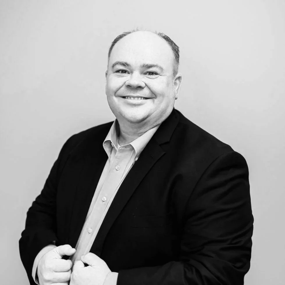 Black and white photo of a smiling man in a suit jacket and dress shirt, standing in front of a plain wall.