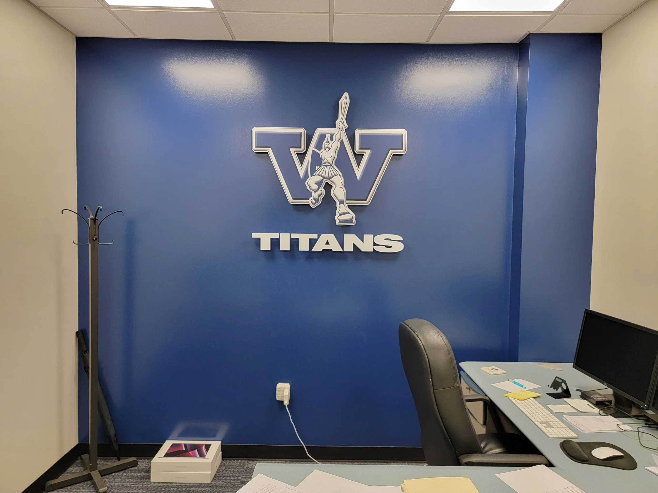 Office corner with a blue wall featuring the Washington Titans logo, a coat rack on the left, a desk with a computer and papers on the right, and a box on the floor.