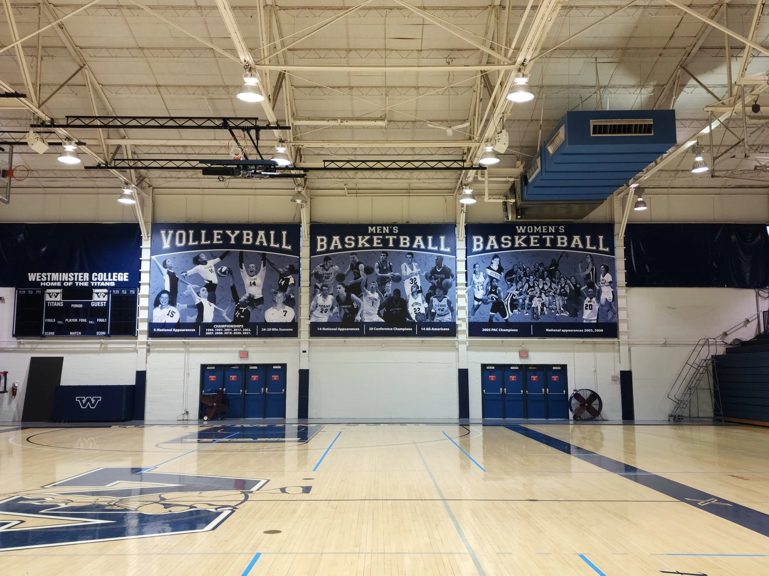 An empty gymnasium with a wooden basketball court, team banners hanging from the wall, and a scoreboard. The gym has bright lights, a basketball hoop, and a large blue ac unit on the ceiling.