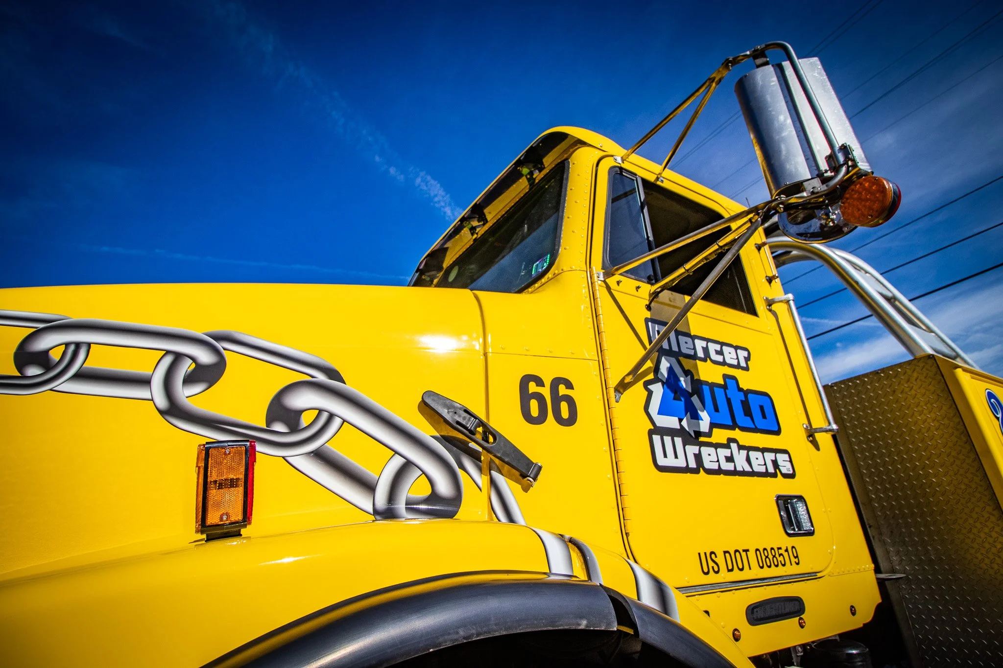 Close-up of a bright yellow tow truck with a chain, a safety light, and a logo that says 'Auto Wreckers' on the side, under a clear blue sky.
