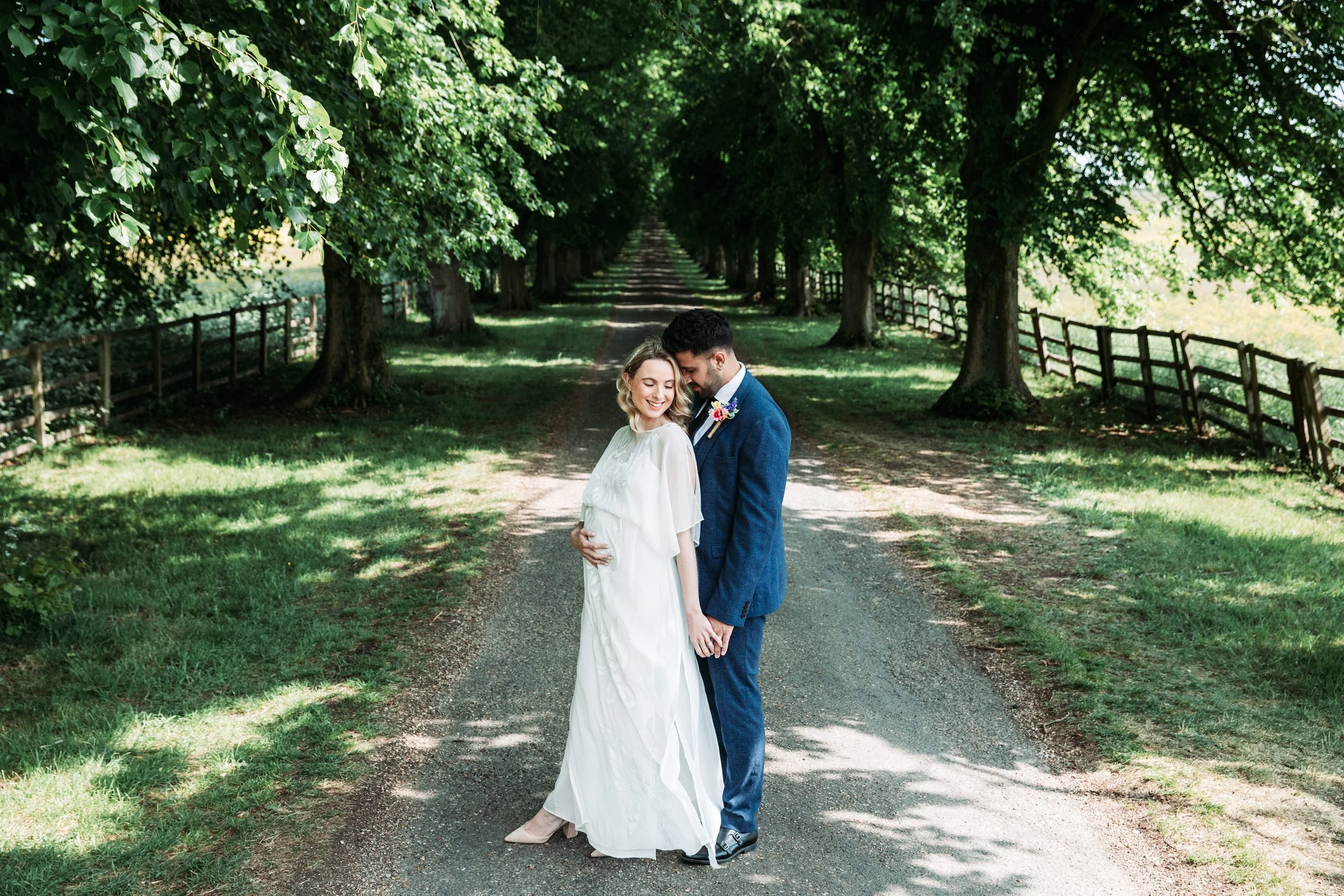 A bride and groom standing on a tree-lined dirt path, smiling and embracing each other, with lush green foliage overhead and wooden fencing on the sides.