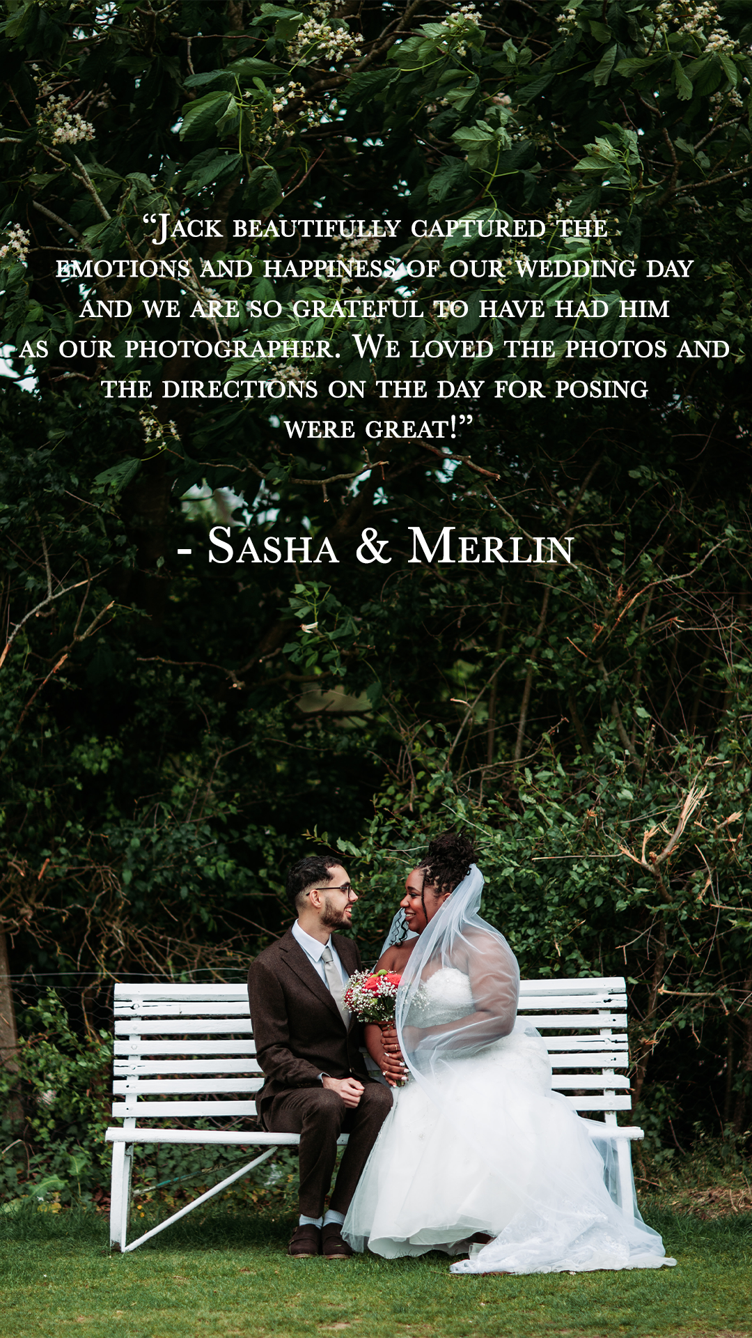 A bride and groom sitting on a white park bench outdoors, smiling at each other, with green foliage and trees in the background.