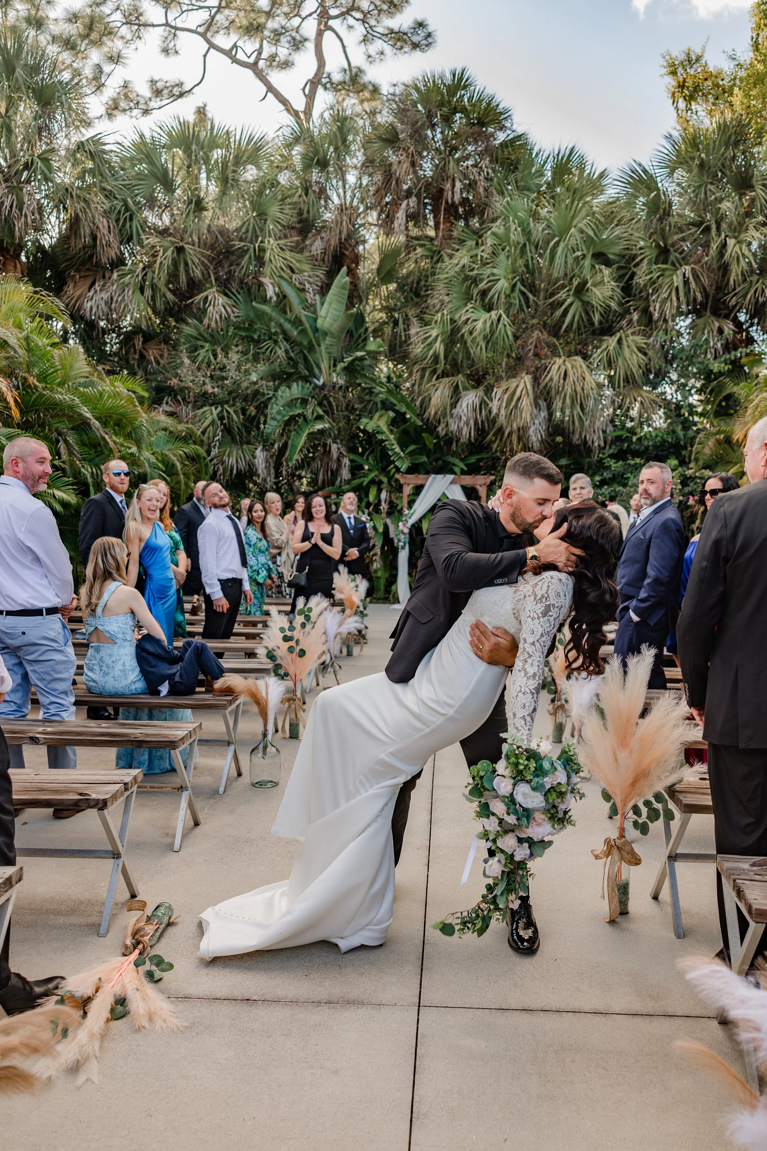 Bride and groom kissing at outdoor wedding ceremony surrounded by guests and decorative floral arrangements with large green plants in the background.