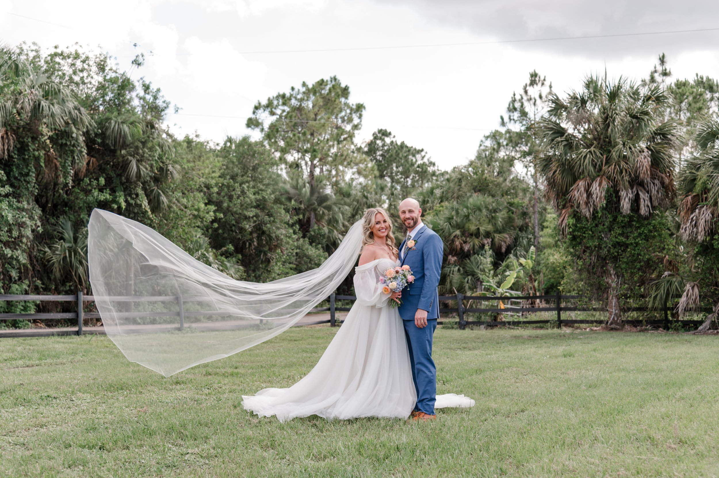 A bride and groom standing together outdoors on a grassy area with lush green trees in the background, the bride wearing a white wedding dress and veil, holding a bouquet of flowers, and the groom in a blue suit.