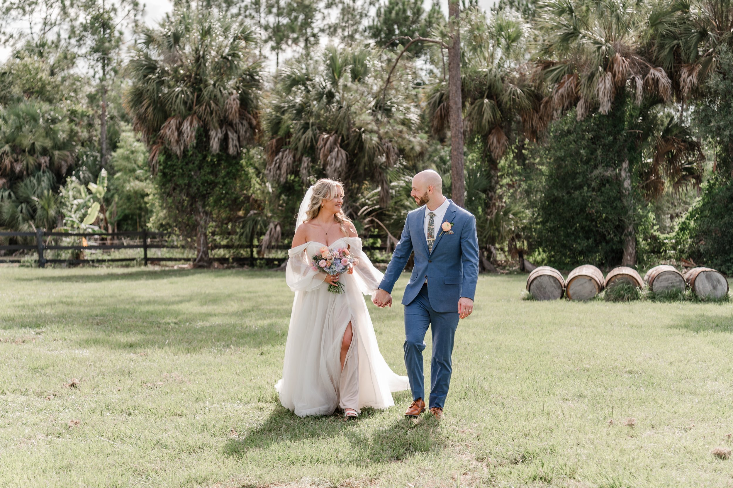 A bride and groom holding hands and walking on green grass outdoors during a wedding ceremony. The bride is wearing a white wedding dress and holding a bouquet, the groom is in a blue suit with a floral tie, and both are smiling.