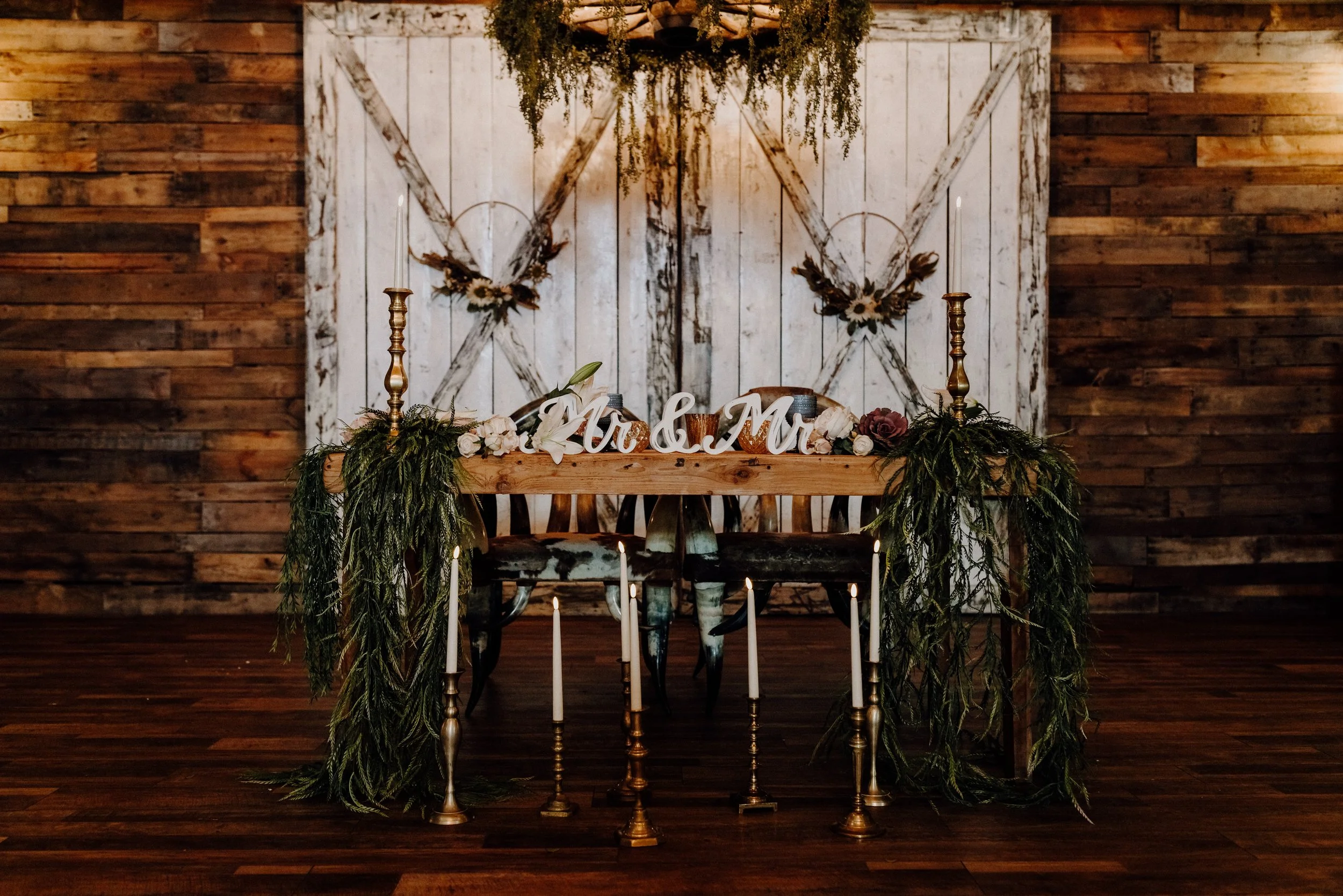 Rustic wedding table setup with "Mr & Mr" sign, wooden table, greenery, candles, and wooden barn doors.
