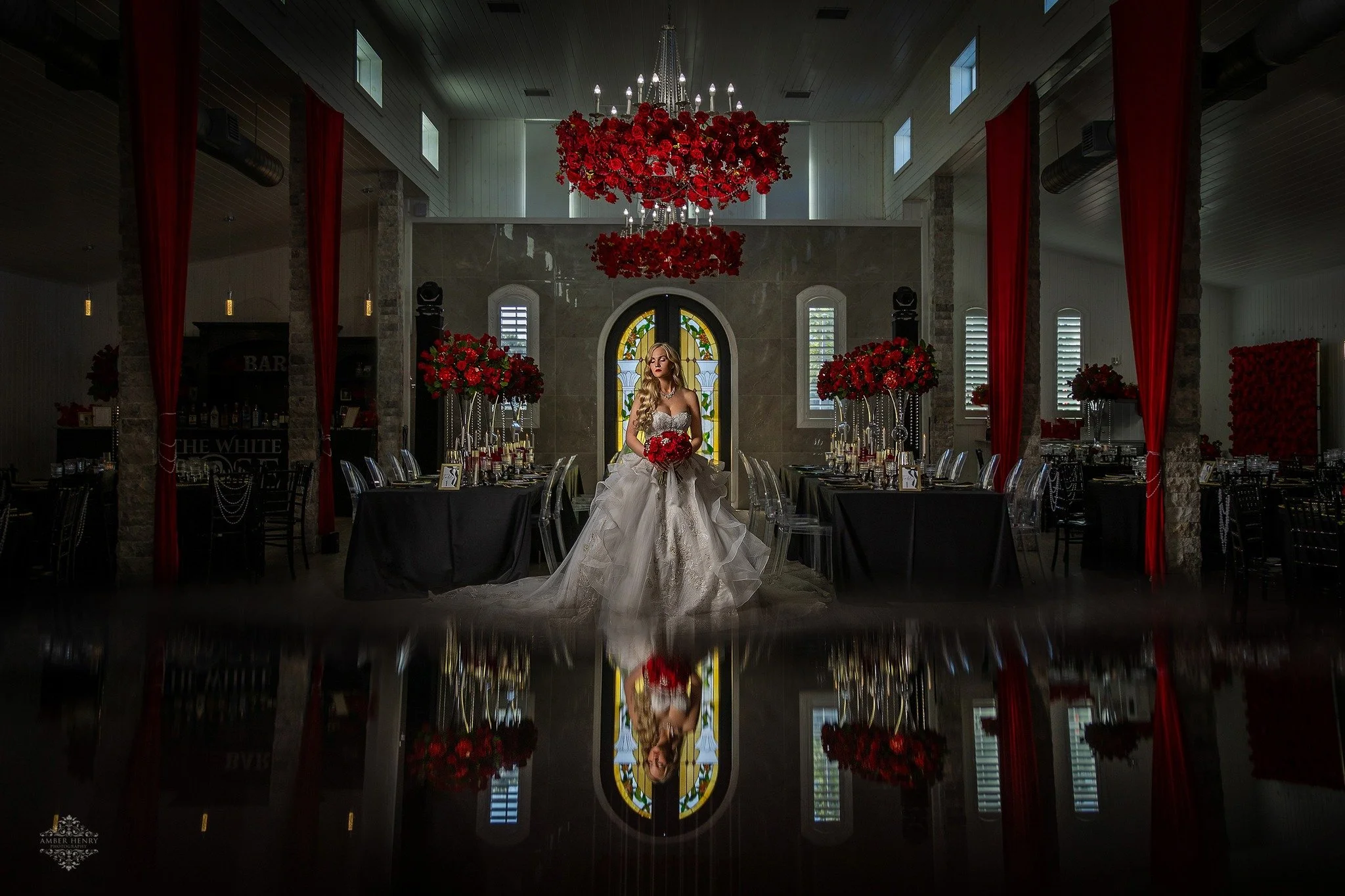 Indoor wedding reception with a bride in a white gown holding a bouquet of red flowers, surrounded by red floral arrangements, chandeliers, red curtains, and stained glass windows.