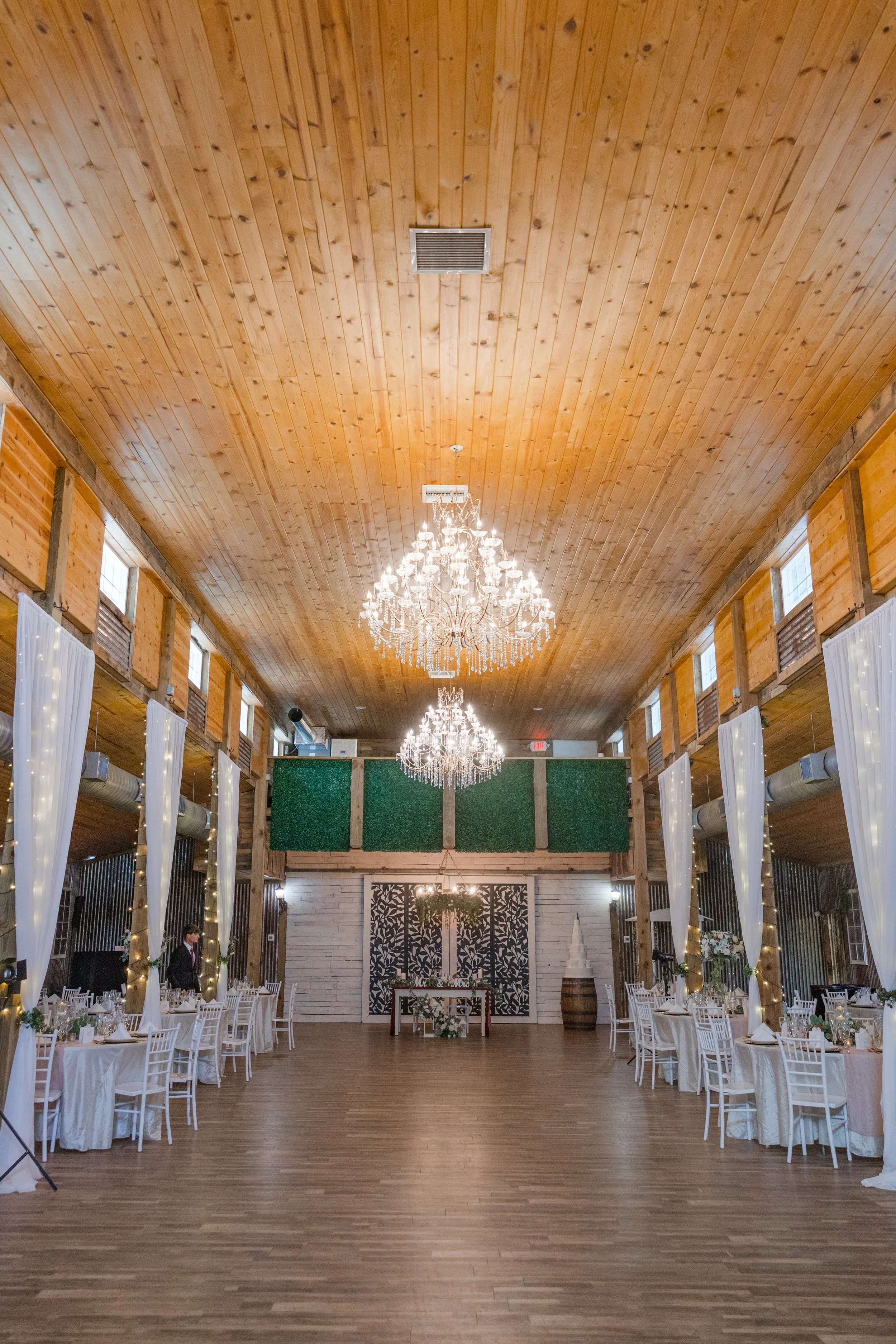 Wedding reception hall with long wooden ceiling, chandeliers, white tables and chairs, decorated for a wedding celebration.