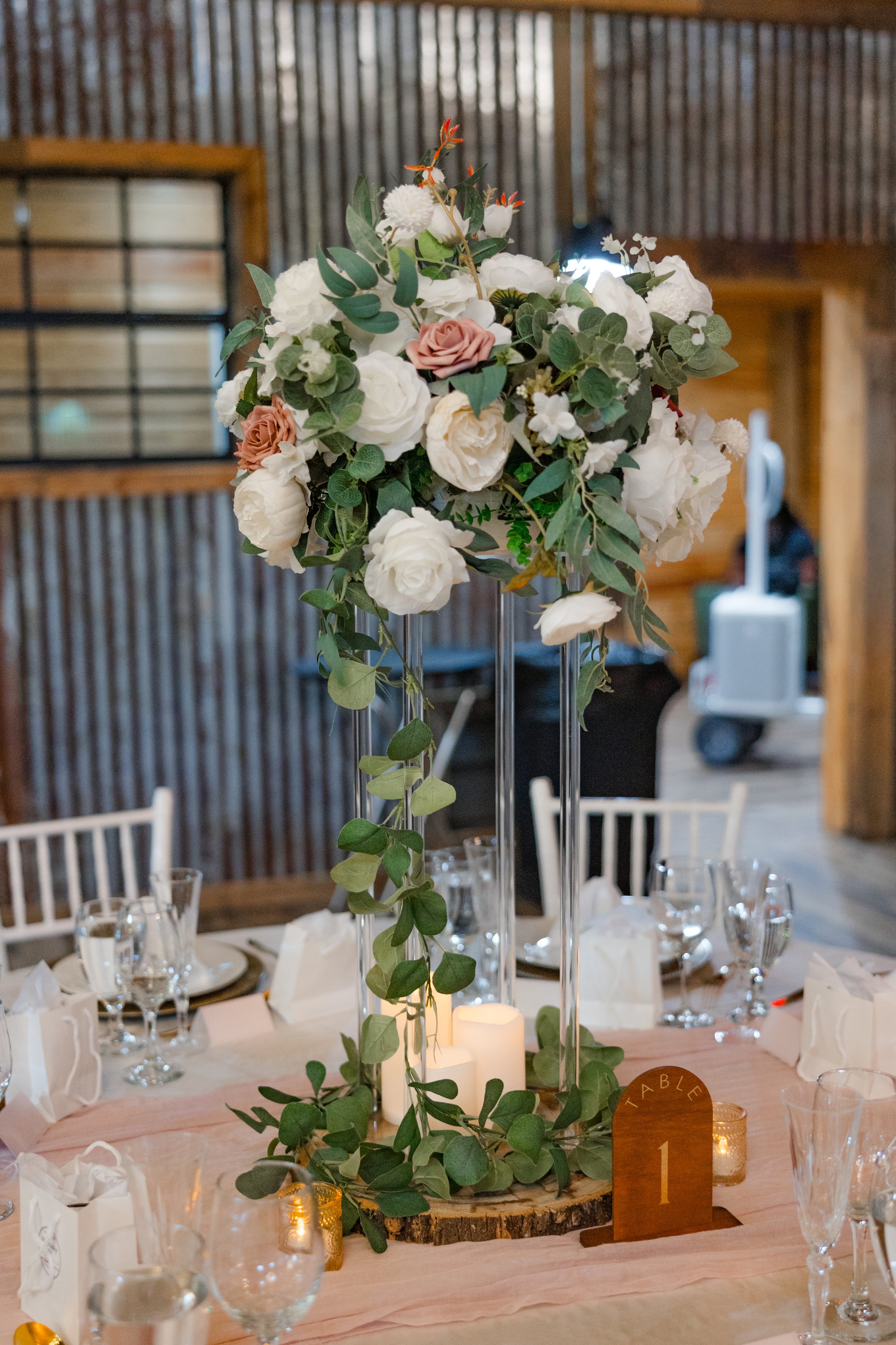 A tall floral centerpiece with white and pink roses, greenery, and small orange accents on a candle-lit table at a rustic indoor wedding reception or banquet setting.