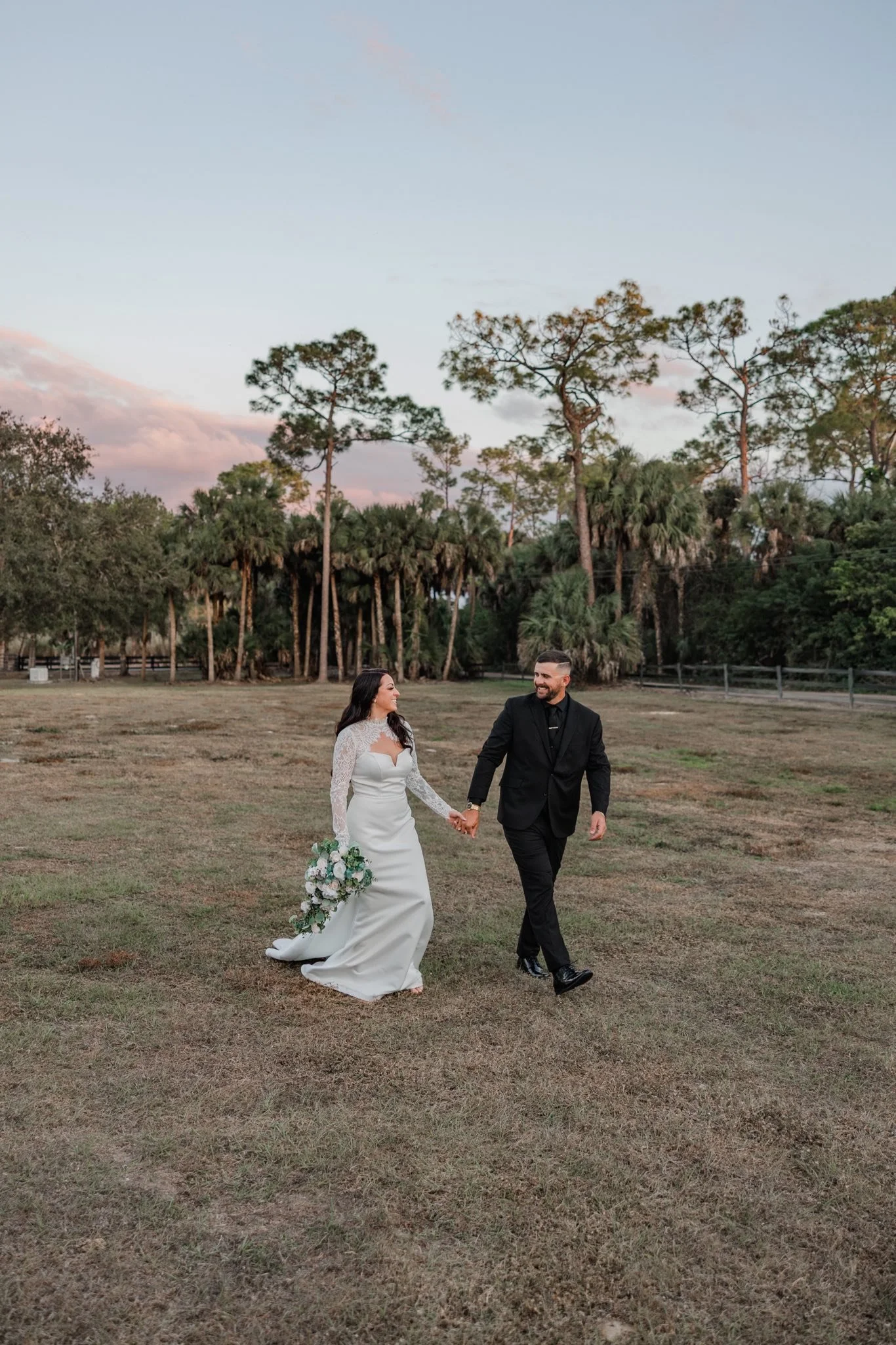A bride and groom holding hands and walking through a grassy field during their outdoor wedding, with trees and a pastel-colored sky in the background.