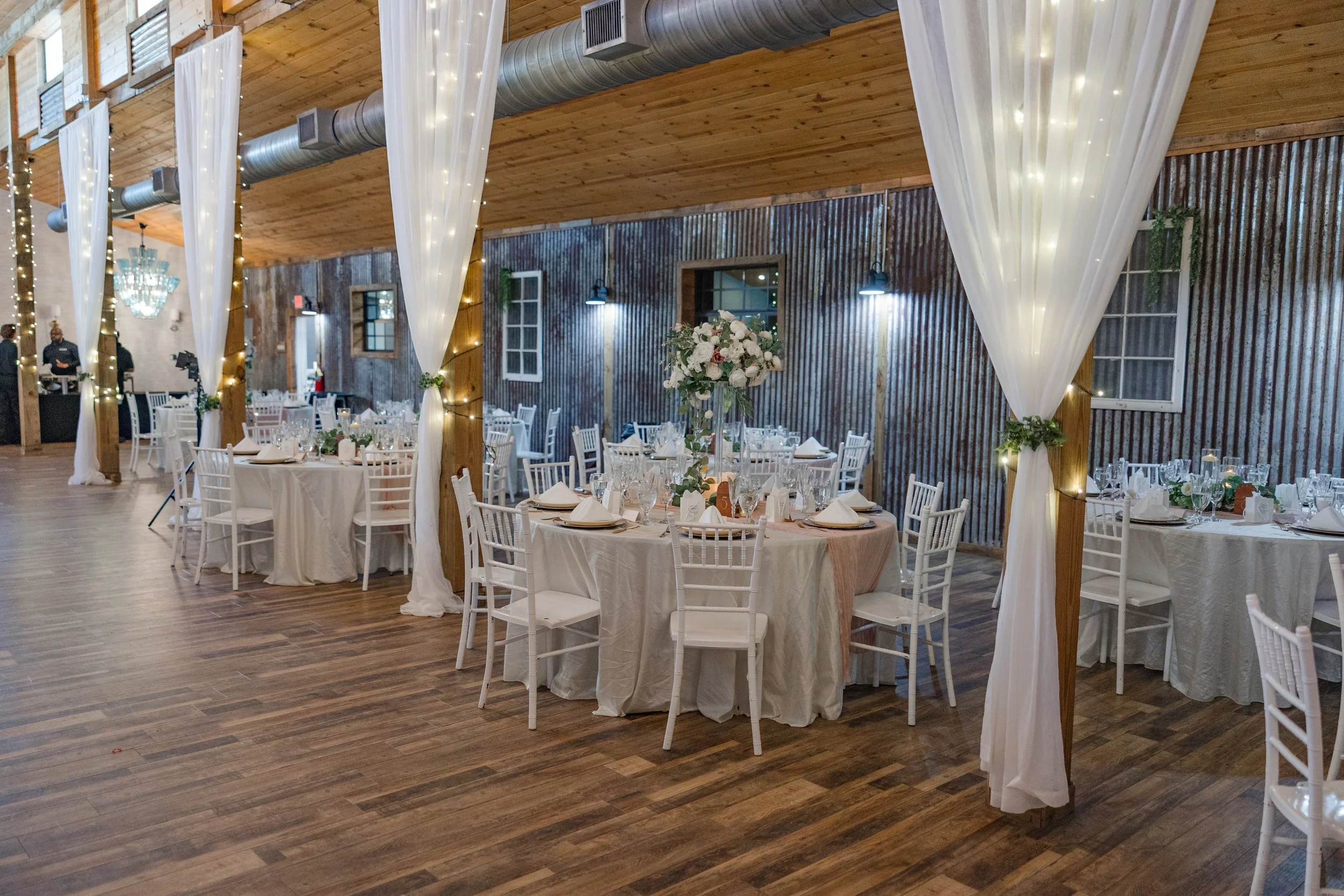 Elegant wedding reception setup in a rustic venue with round tables draped in white tablecloths, white chairs, floral centerpieces, and white drape curtains with fairy lights hanging from the ceiling, wooden walls, and hardwood floors.