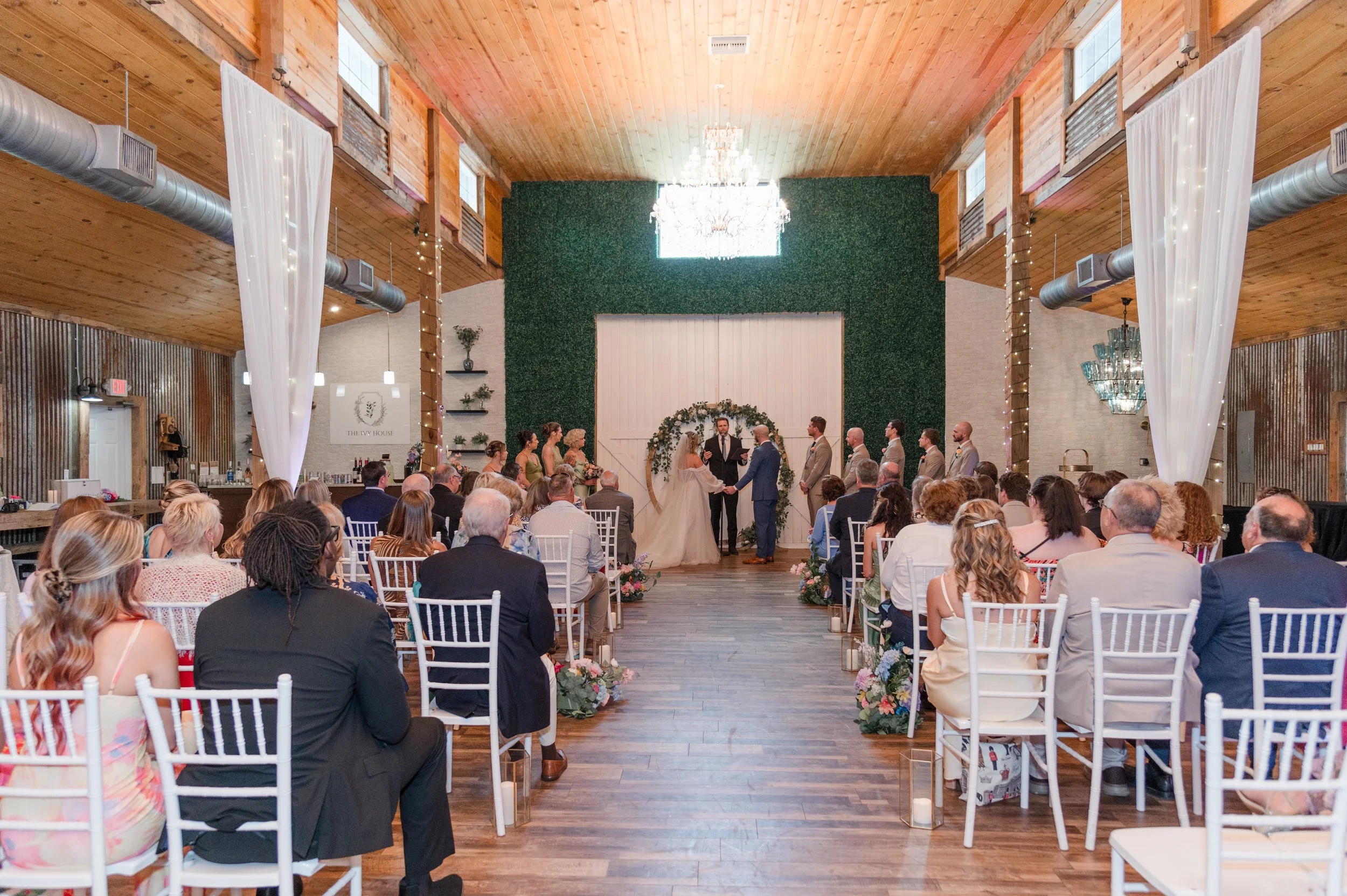 Indoor wedding ceremony with bride and groom exchanging vows in front of officiant, guests seated on white chairs, decorated with flowers and greenery, wooden interior with high ceiling, chandeliers, white curtains, and fairy lights.