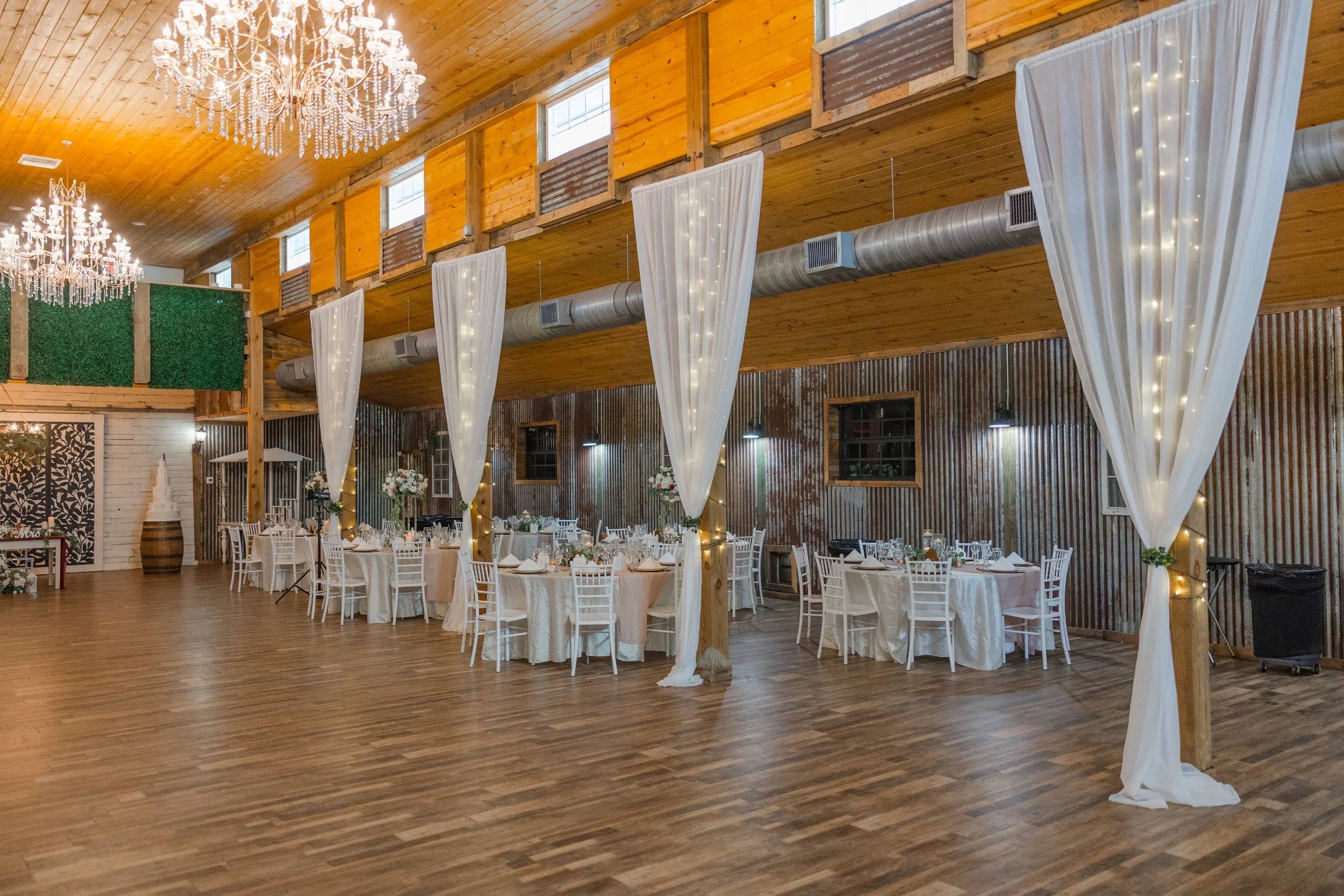 A rustic wedding or event venue decorated with chandeliers, white drapes with twinkle lights, banquet tables with white tablecloths, floral centerpieces, and white chairs. The venue features a wood-paneled ceiling, exposed ductwork, and a corrugated 