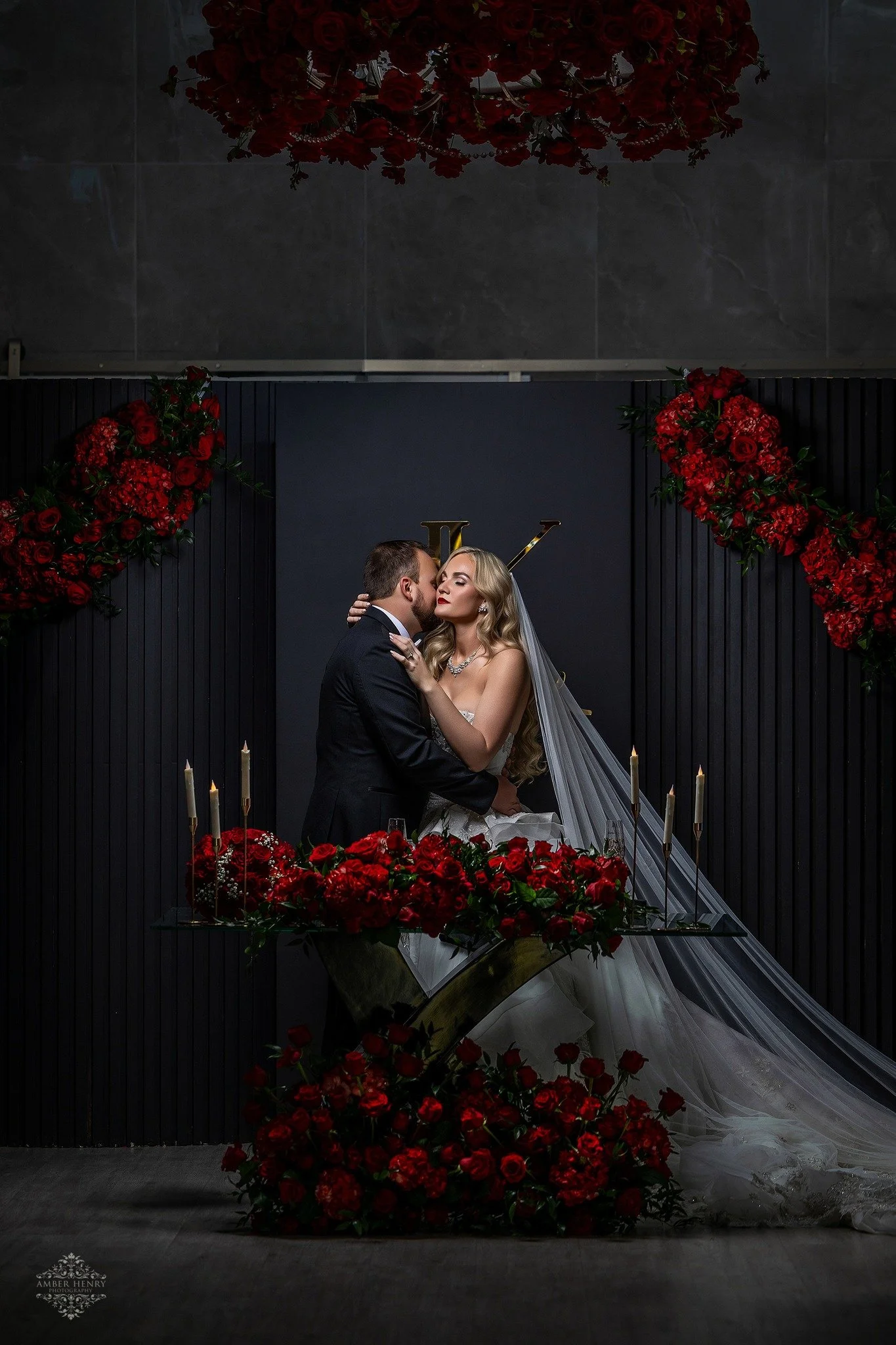 A bride and groom sharing a kiss at their wedding ceremony, with the bride wearing a white wedding gown and veil, surrounded by red roses and candles, against a dark backdrop with the initials LV.