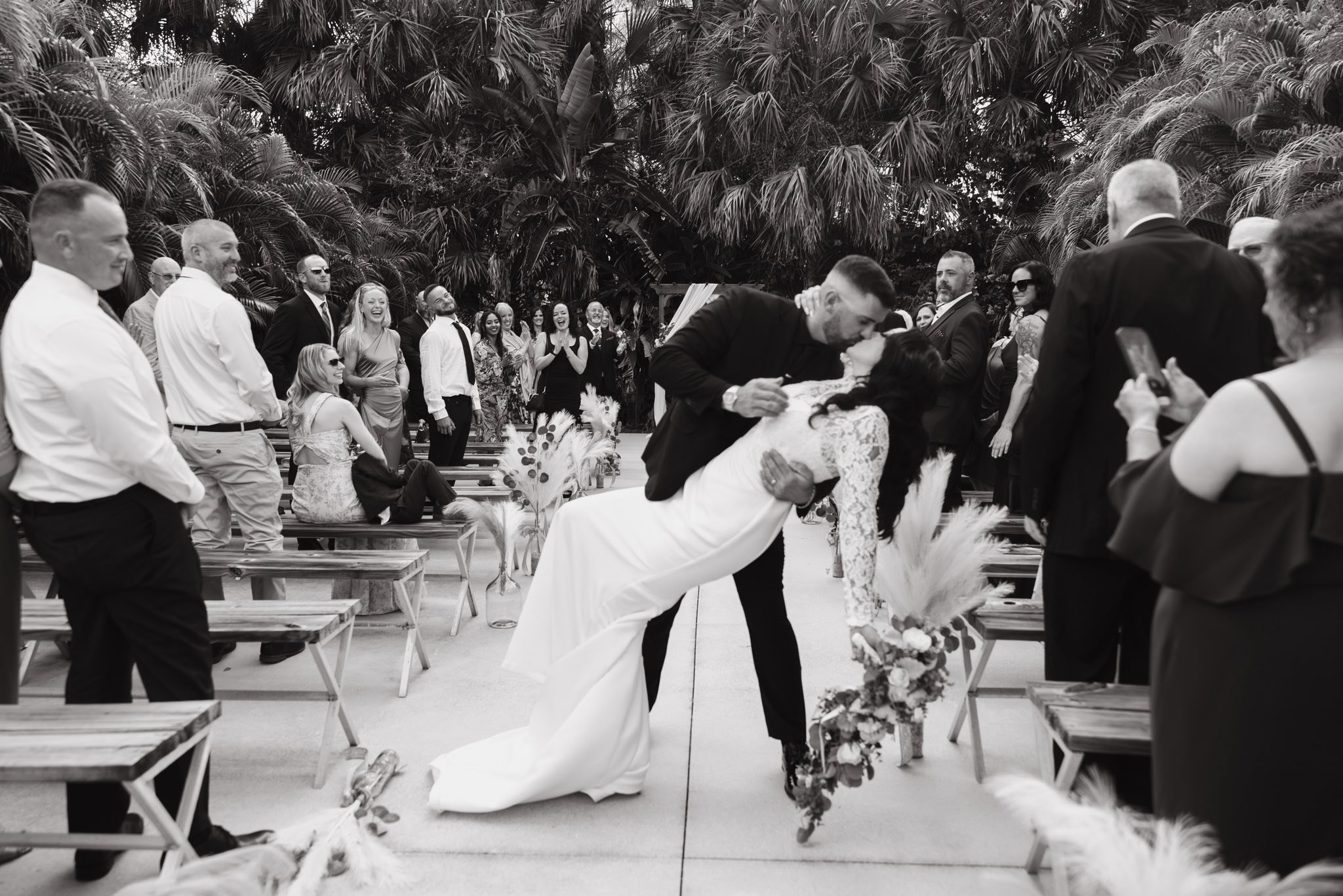 A wedding ceremony with guests watching as the bride and groom kiss, outdoors with lush tropical trees in the background.