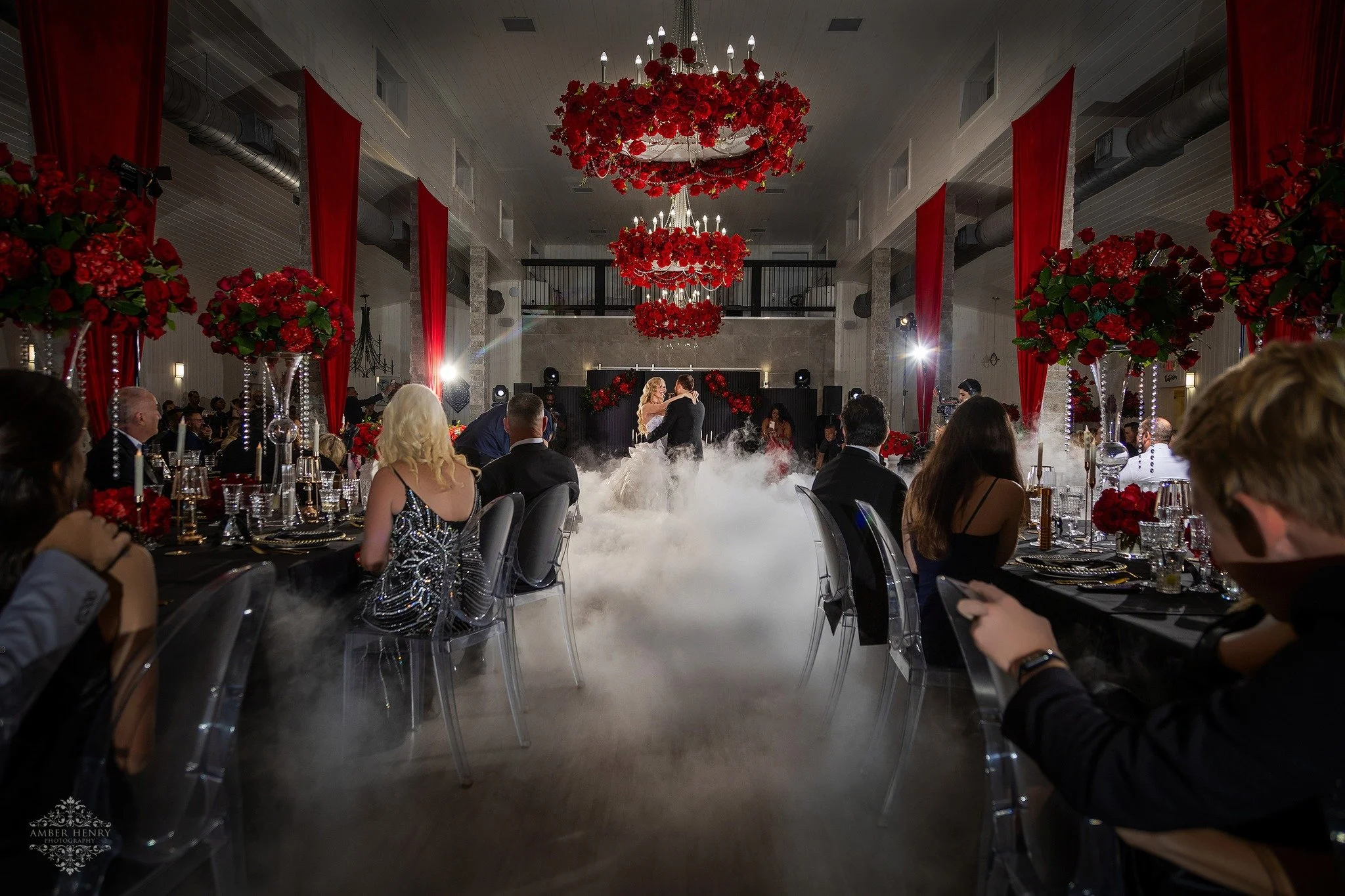 A wedding reception with a bride and groom dancing on a foggy floor, surrounded by seated guests in a decorated hall with red floral arrangements and red curtains.