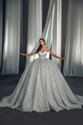 A woman in a strapless, white wedding gown with a full, glittery skirt standing with arms outstretched in front of floor-to-ceiling curtains.