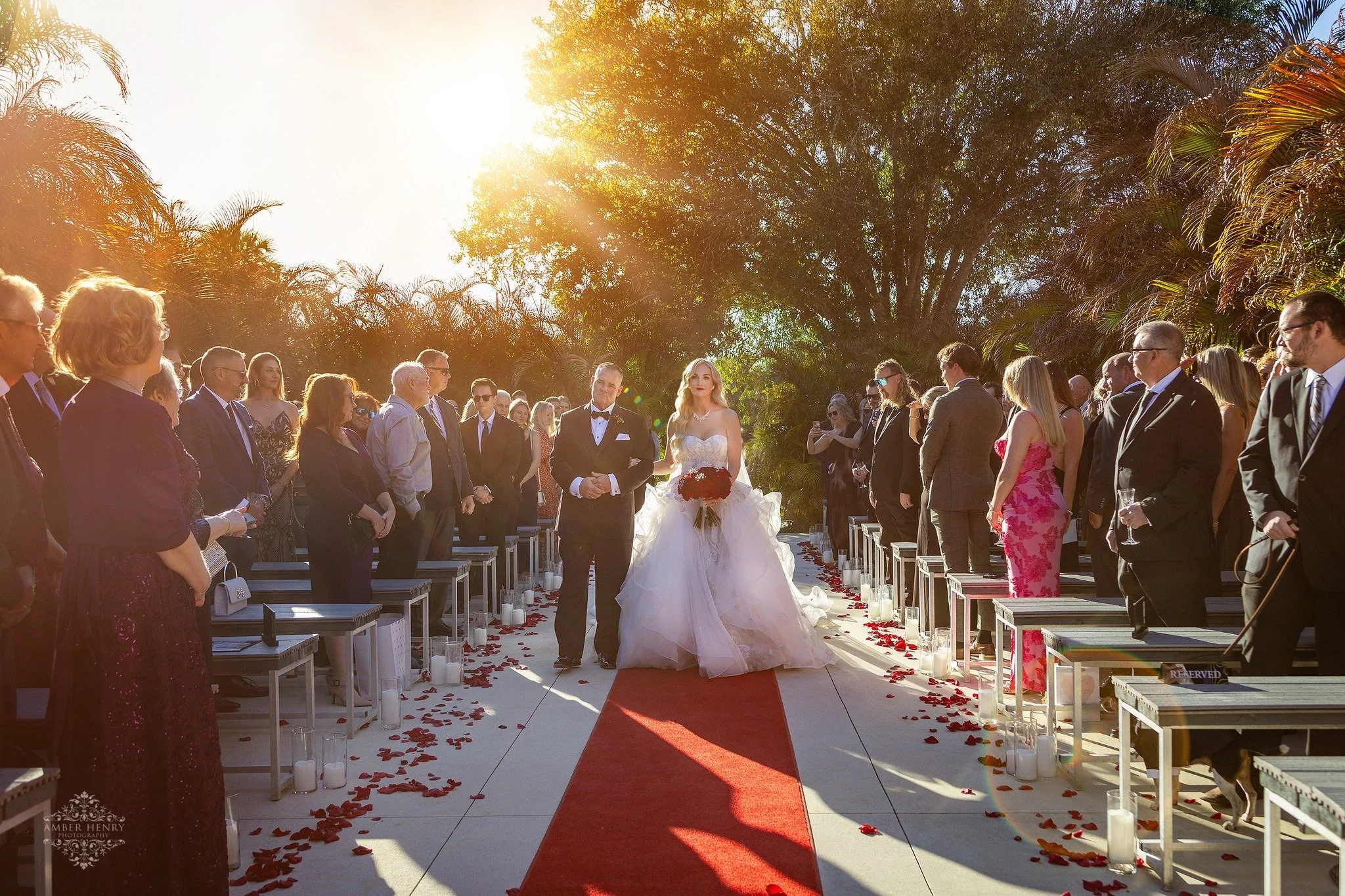A bride in a white gown walks down an aisle lined with rose petals, escorted by a man, with guests seated on either side inside a decorated wedding venue.