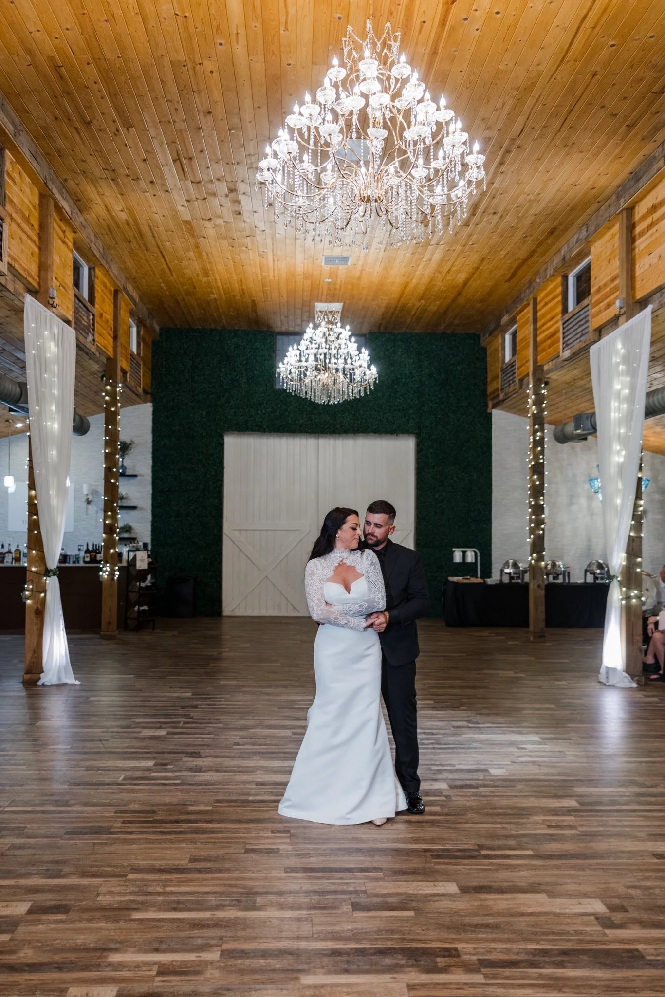 A bride and groom are dancing closely in a rustic wedding venue with wooden ceilings, chandeliers, and string lights, creating a romantic atmosphere.