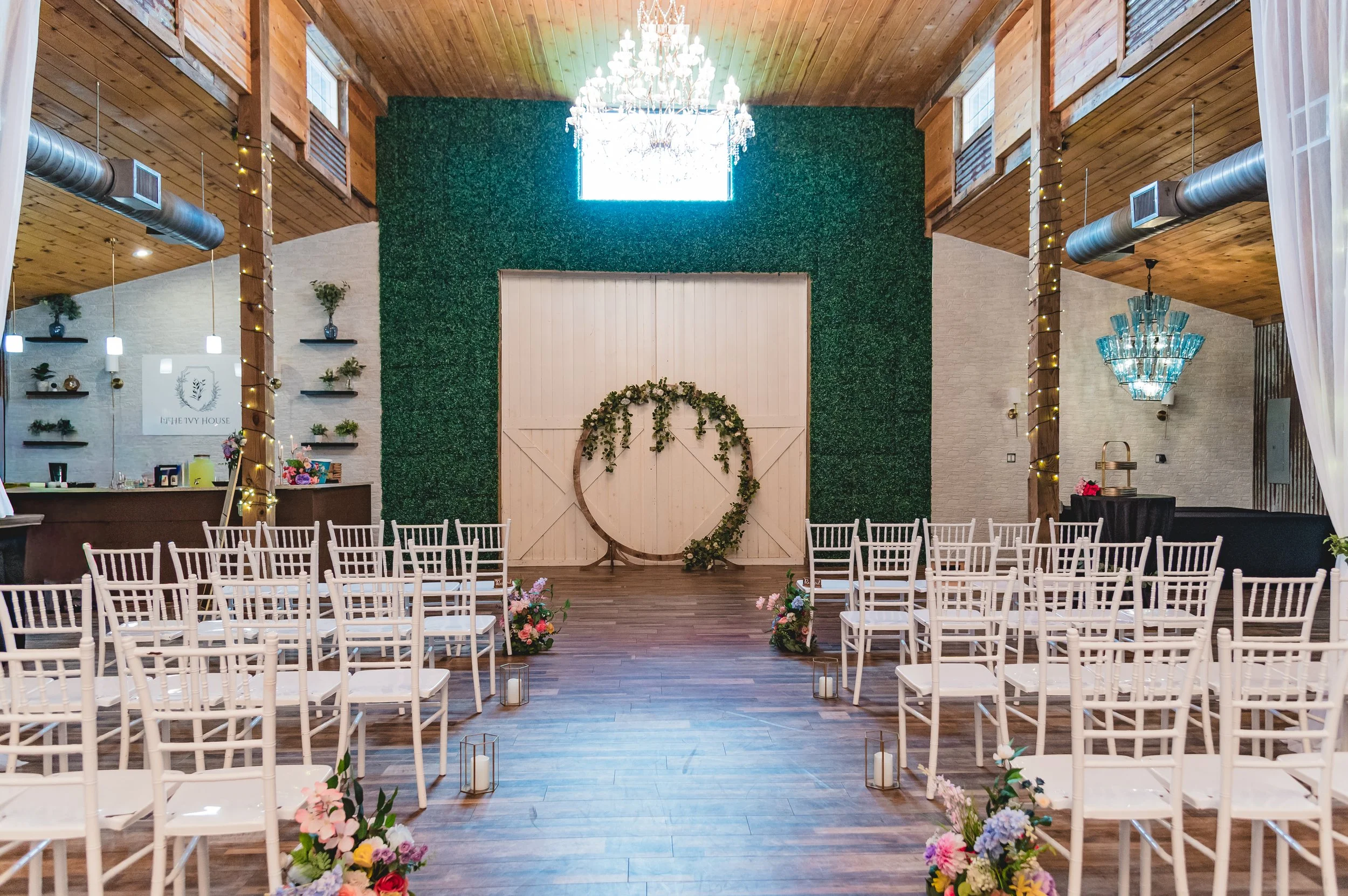 Indoor wedding or event space with rows of white chairs facing a decorated wall with floral arrangements, large circular behind the chairs, white candles, green foliage, and a chandelier hanging from the ceiling.