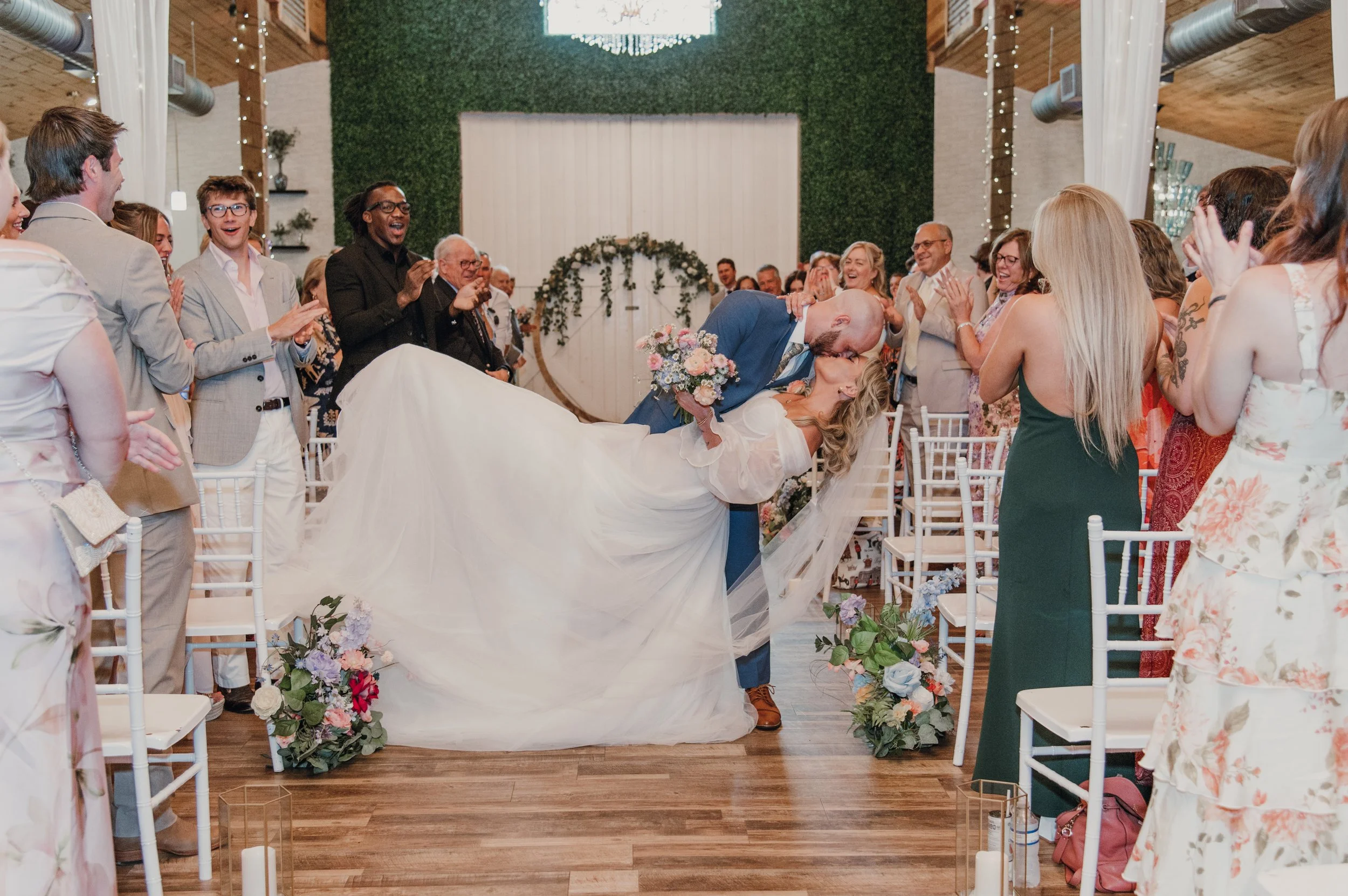 A wedding celebration with the bride and groom sharing a kiss while the groom holds a bouquet, surrounded by guests clapping and smiling in a decorated indoor venue.