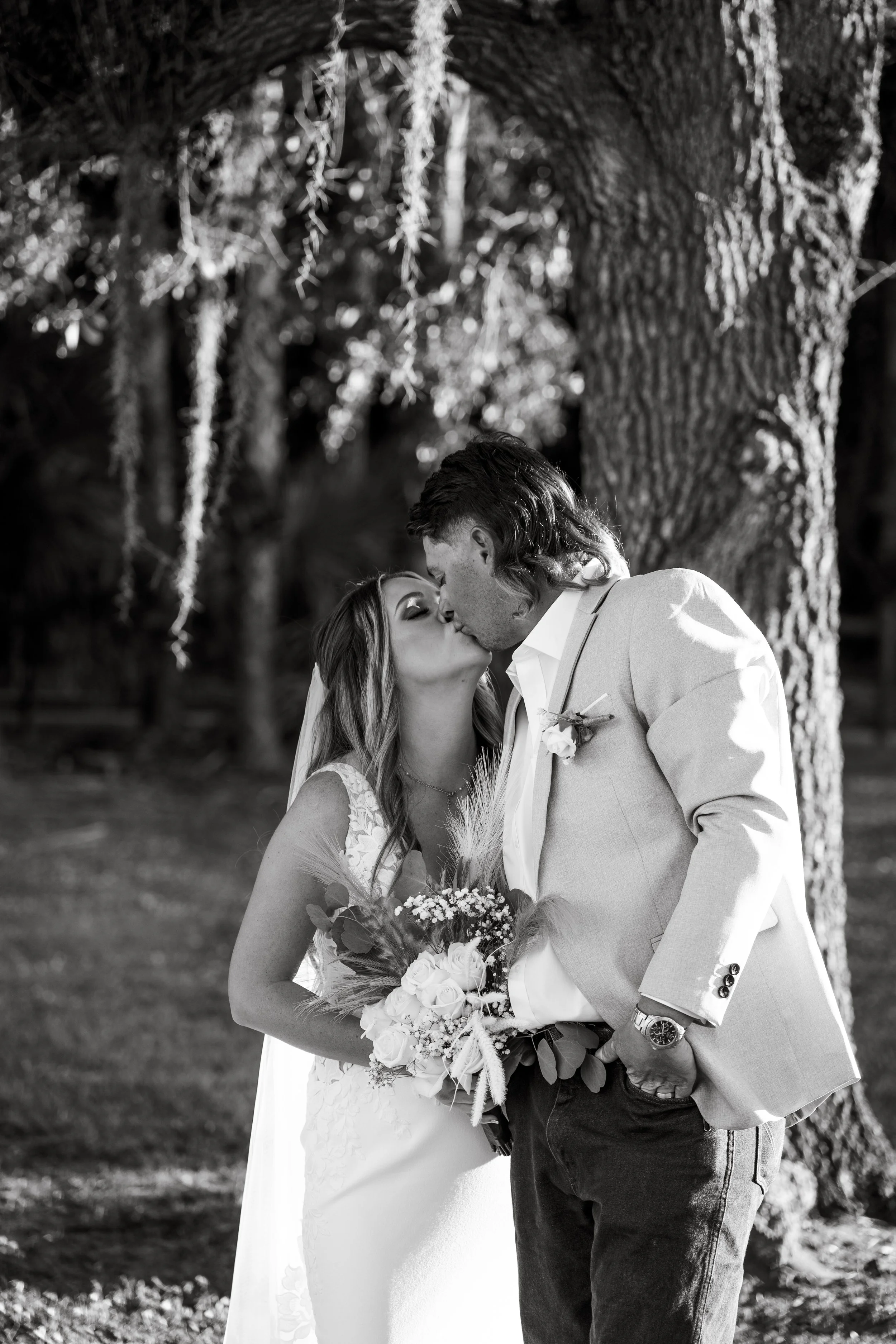 Bride and groom celebrating at their wedding, holding hands triumphantly. The bride is in a lace wedding dress holding a bouquet, and the groom is in a beige suit with a bow tie.