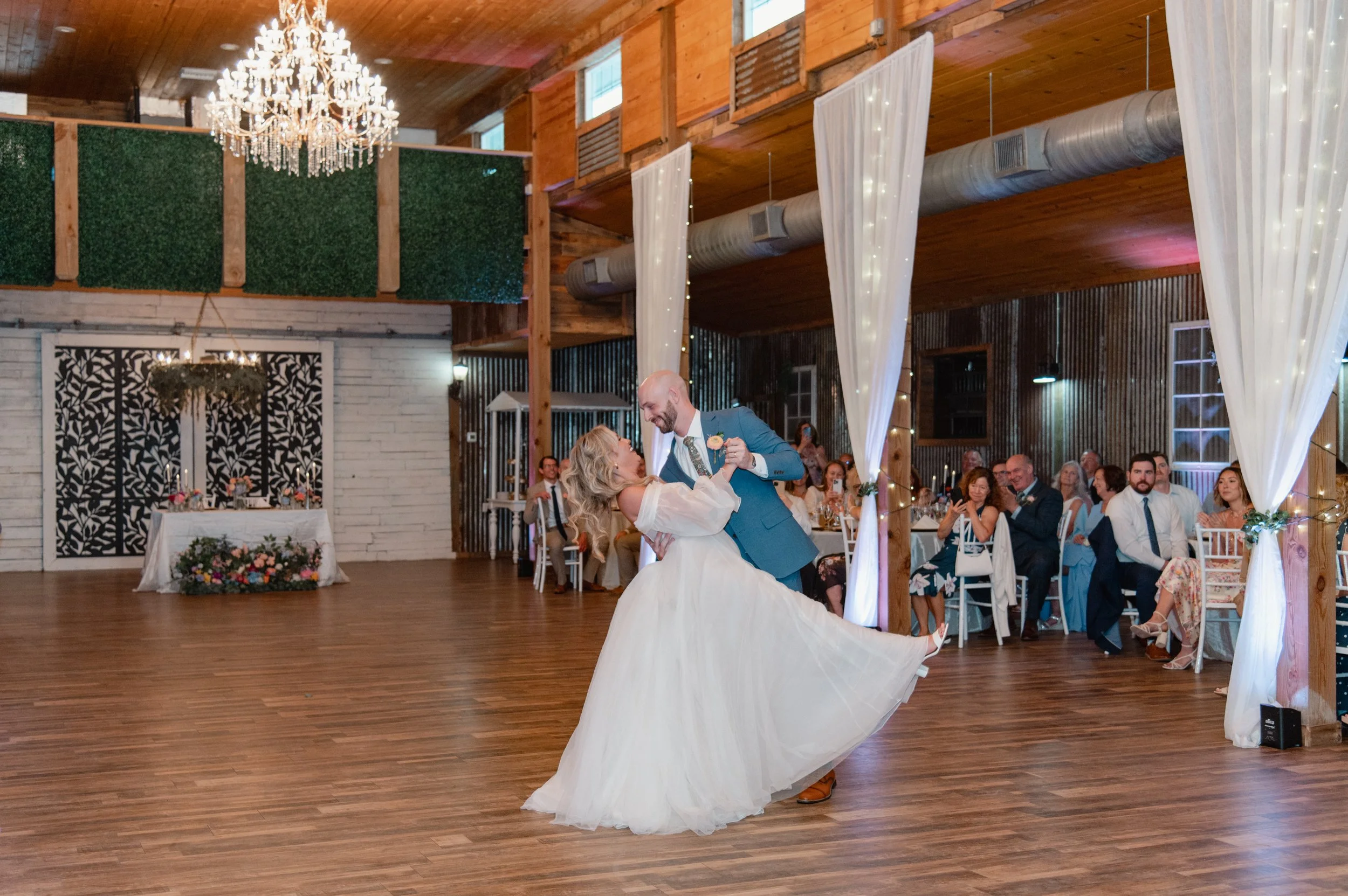 A bride and groom dancing during their wedding reception in a rustic barn-style venue with wood walls and ceiling, a large chandelier, white drapes, and guests seated at tables watching the couple.
