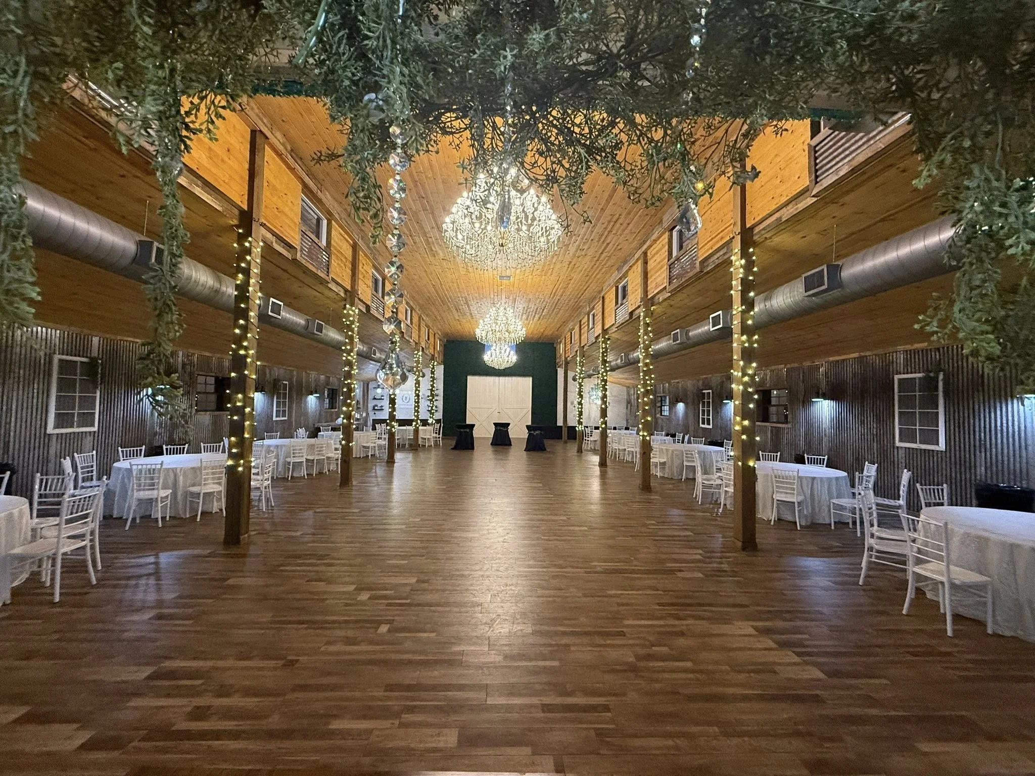 Wedding reception hall with draped white fabric and string lights, wooden walls and ceiling, round tables with white tablecloths and chairs, and a decorated head table at the front.