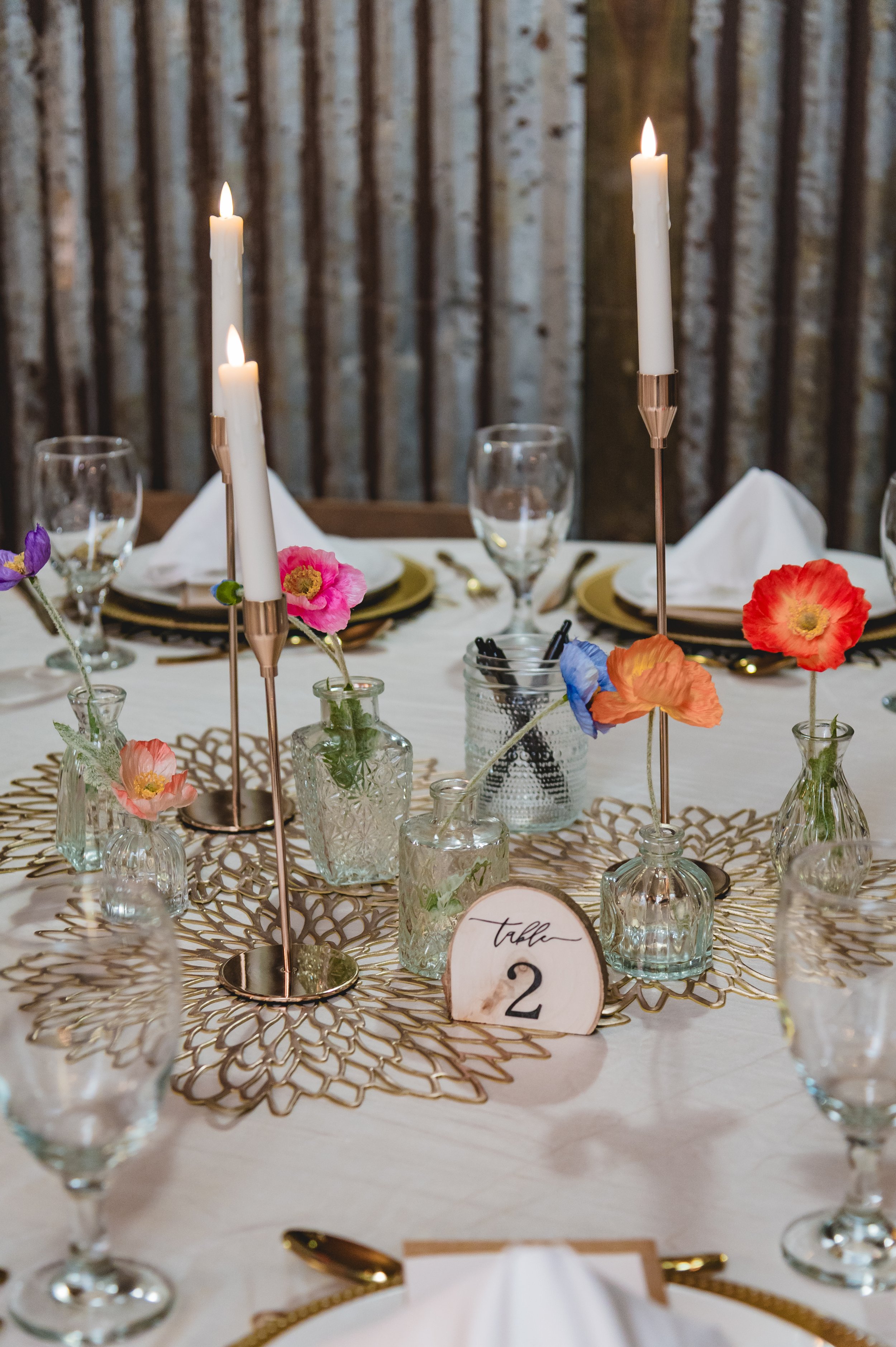 A wedding or formal event table setting with tall white candles in brass holders, colorful flowers in small glass vases, place cards, glassware, and gold cutlery on a white tablecloth with a decorative gold placemat, in front of a wooden tree backgro