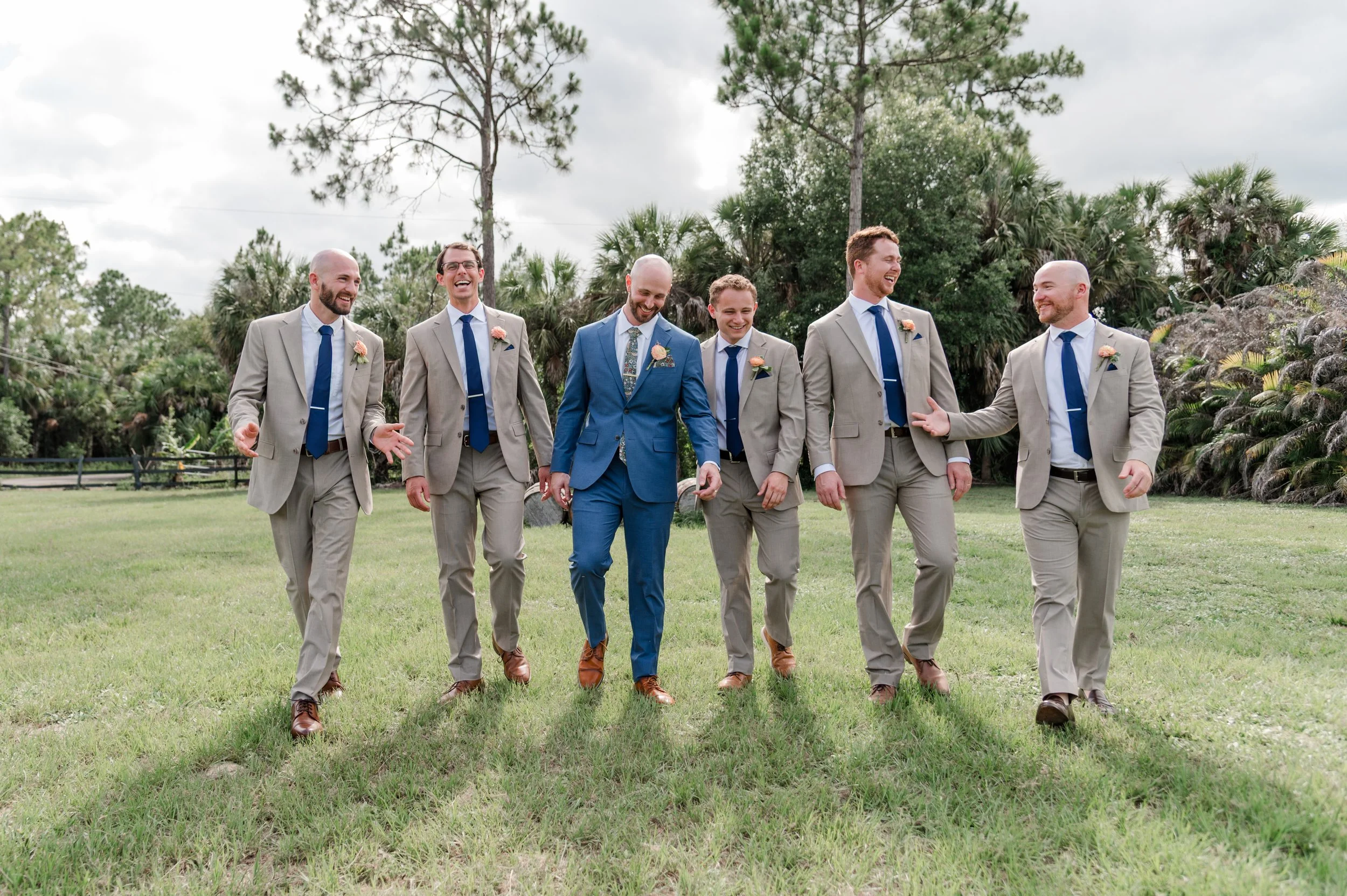 A group of seven men dressed in suits, walking on a grassy field outdoors, surrounded by trees, and smiling and laughing.