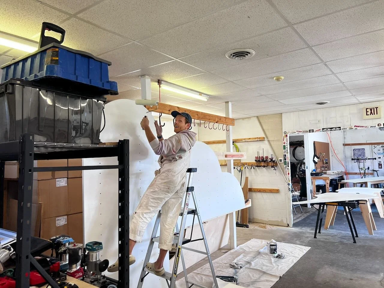 A man standing on a ladder working on a large structure inside a workshop. The workshop has various tools, shelves, and workbenches, with an exit sign visible in the background.