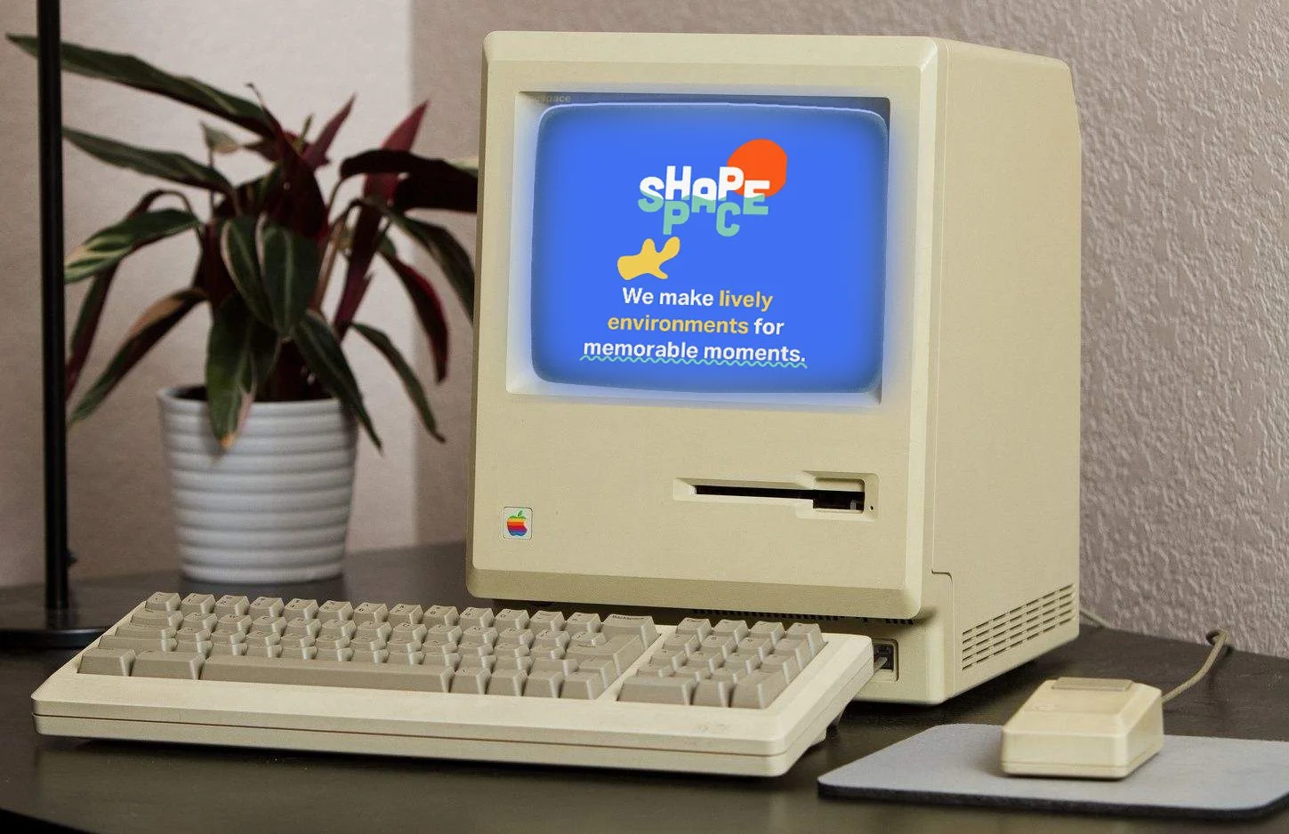 A vintage Apple Macintosh computer with a small screen displaying a colorful logo and text. The computer has a beige keyboard and a mouse, both matching the color of the computer. A potted plant with long green and red leaves is in the background.