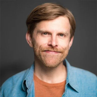 Portrait of a man with light brown hair and beard, wearing a blue shirt over an orange T-shirt, against a gray background.