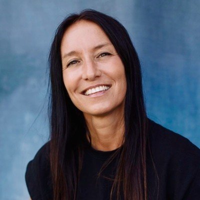 A woman with long dark hair smiling, wearing a black shirt, standing outdoors against a blue background.