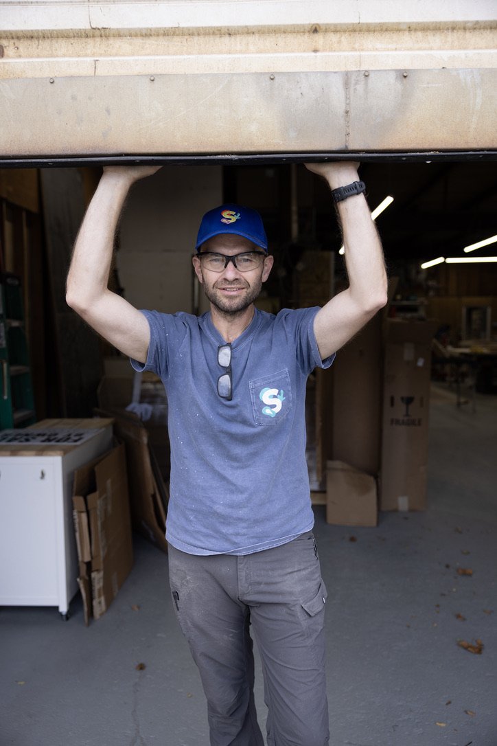 A man in a blue cap, glasses, and gray t-shirt holding up a large wooden object in a workshop.