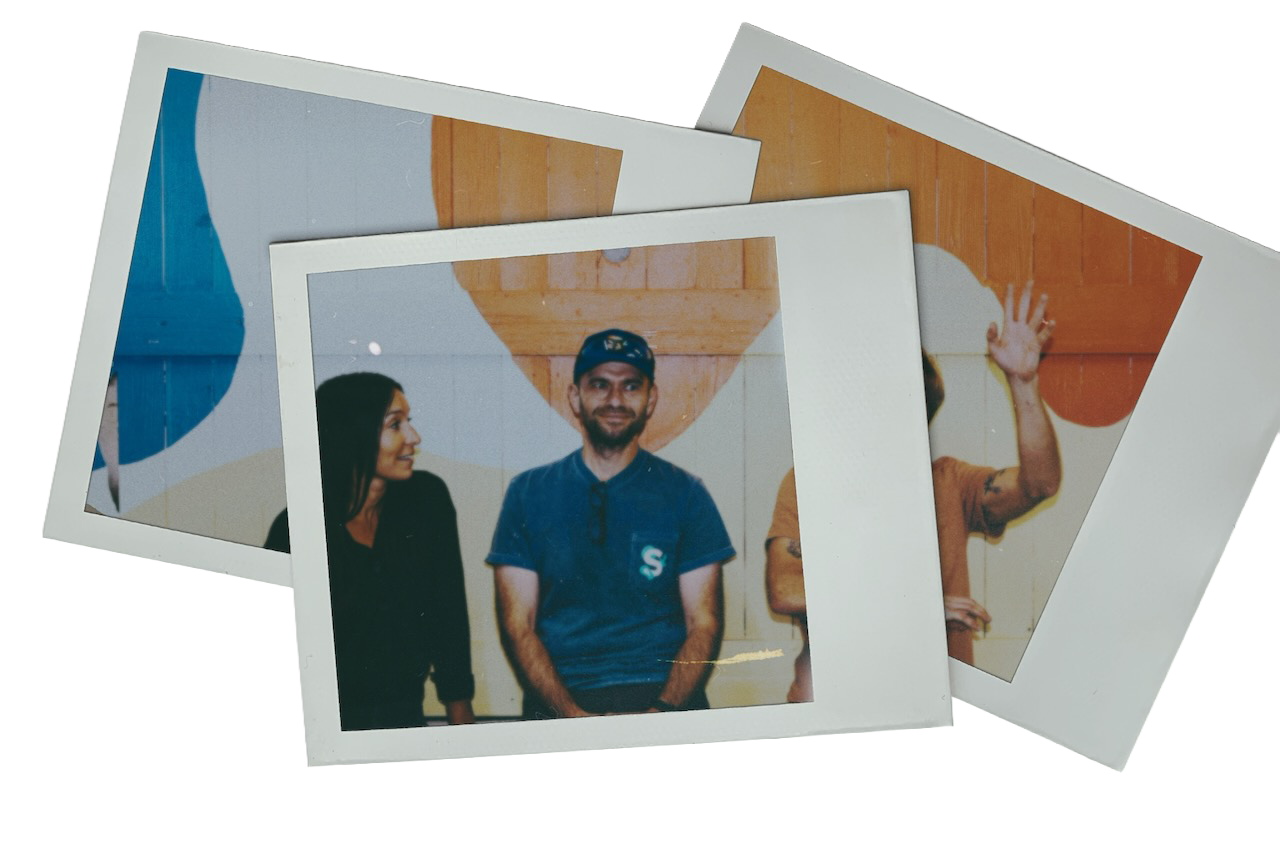 Three people standing and posing for photographs against a colorful wall with abstract shapes, with one person waving.