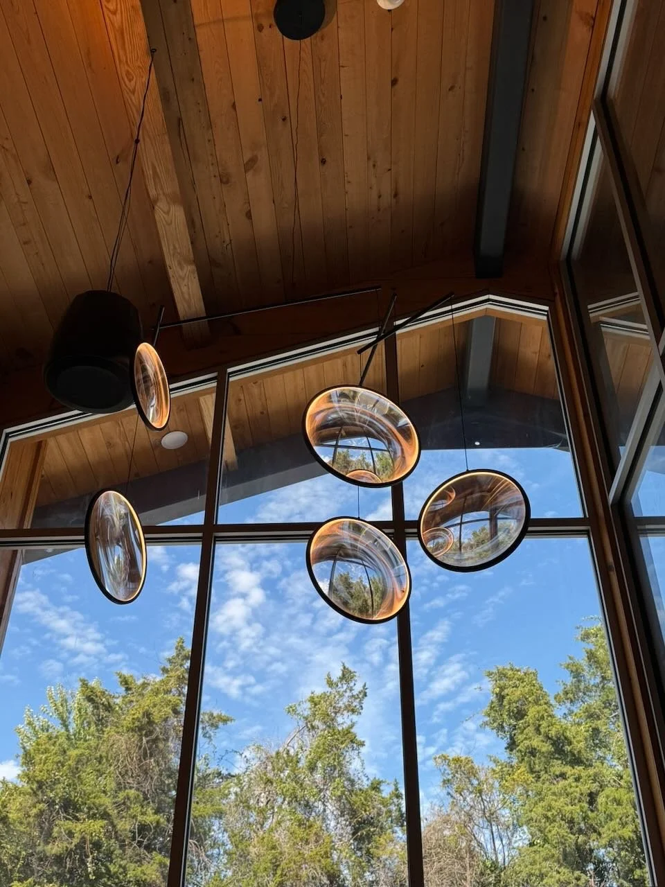 Interior view of a room with high large glass windows showing a blue sky with scattered clouds and green trees outside, wood-paneled ceiling, and modern hanging pendant lights.