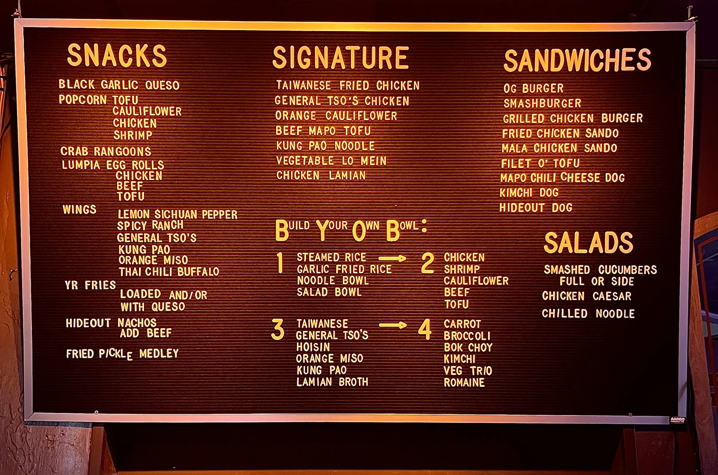 A restaurant menu board with sections for Snacks, Signature dishes, Sandwiches, and Salads written in yellow text on a black background with red border, listing various Asian-inspired food options.