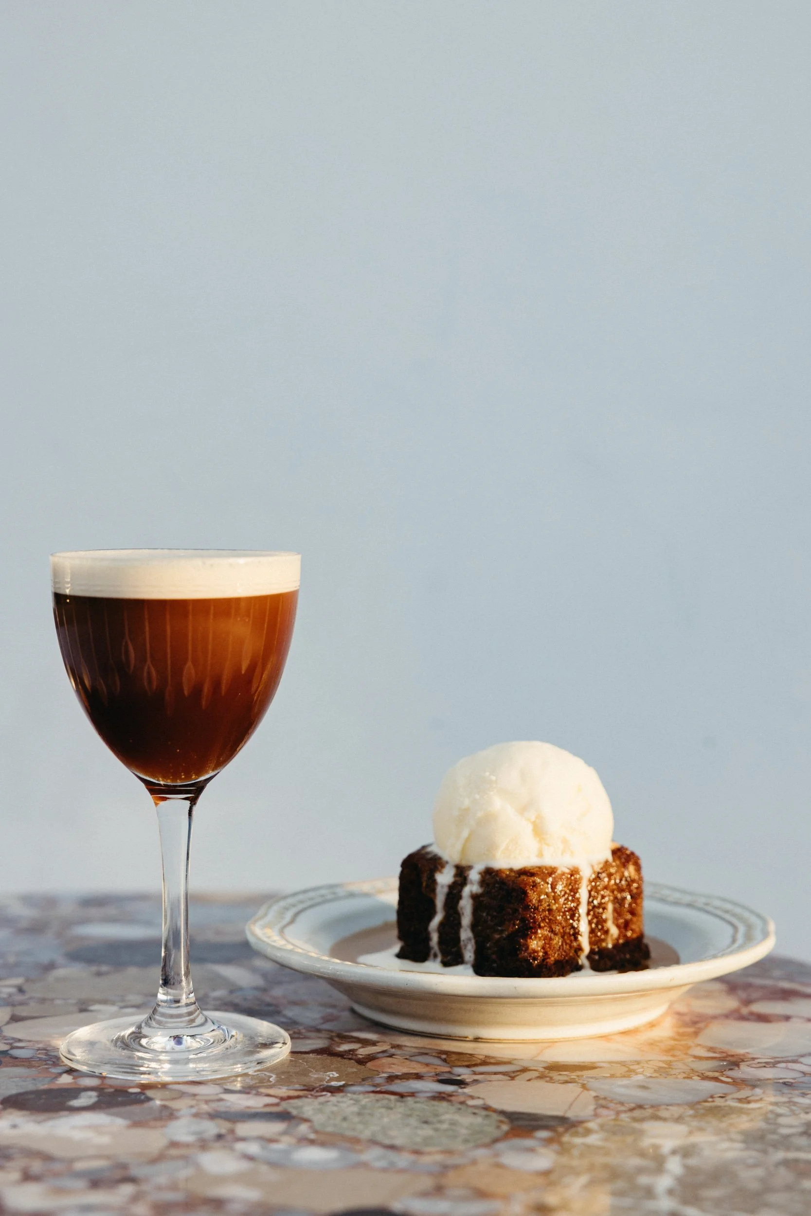 Espresso martini, a date cake with vanilla ice cream on top, on a marble table against a light blue sky background.
