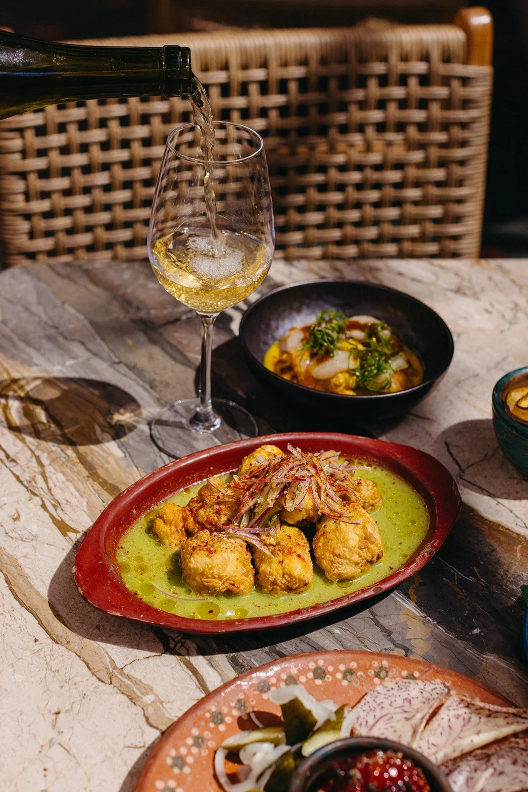 A glass of white wine being poured at a table with various dishes, including a plate of fried food in a yellow sauce, a bowl of seafood, and a plate of sliced vegetables, on a marble table with a woven chair in the background.