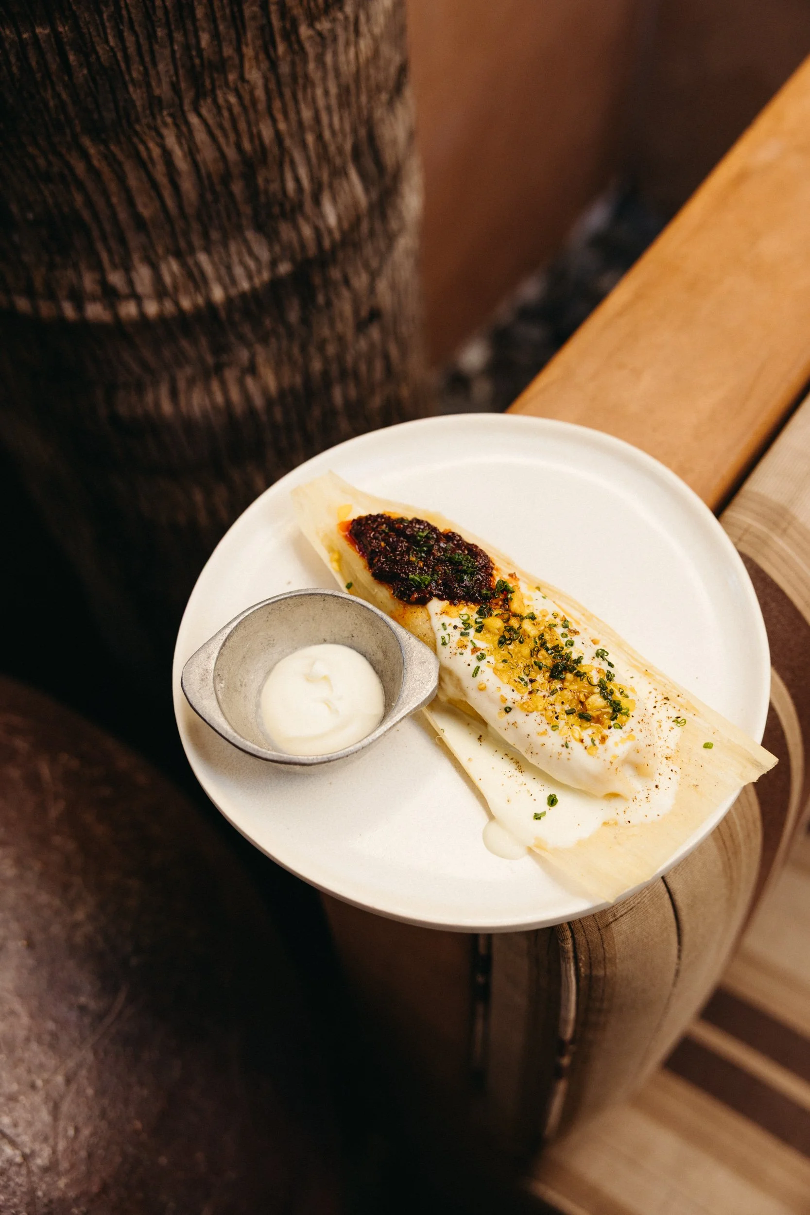 A sweet corn bollo, topped with sauce and garnished with herbs and spices, accompanied by a small container of white sauce, on a wooden table.